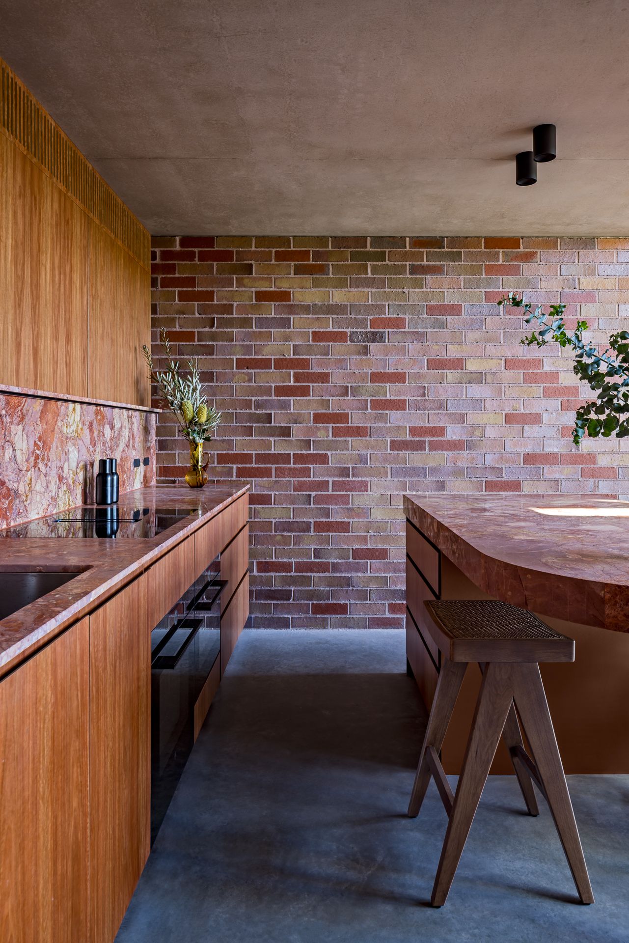 Modern kitchen with timber cabinetry, marble island, wooden stool, brick wall and polished concrete floor under a concrete ceiling.