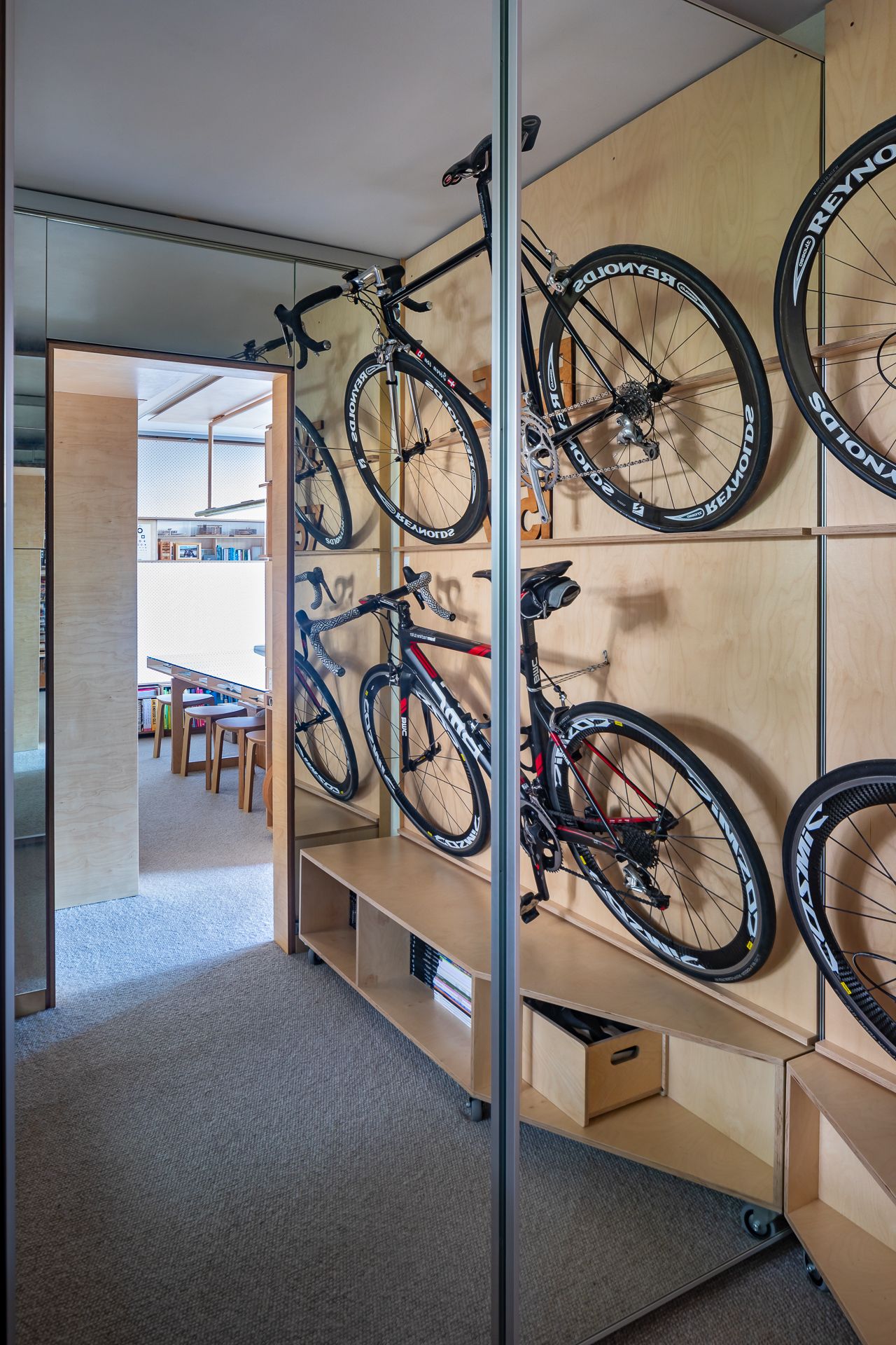 Two road bicycles hang on a plywood wall with rolling shelves, while a mirrored door reflects a study space with stools and a long desk.