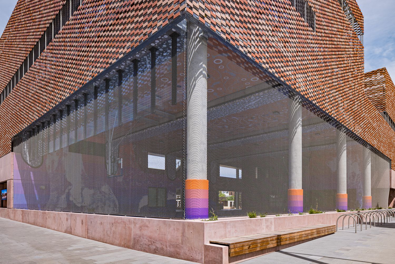 Modern brick building with a mesh-draped glass corner, concrete columns with colorful striped bases, and a wooden bench along the sidewalk.
