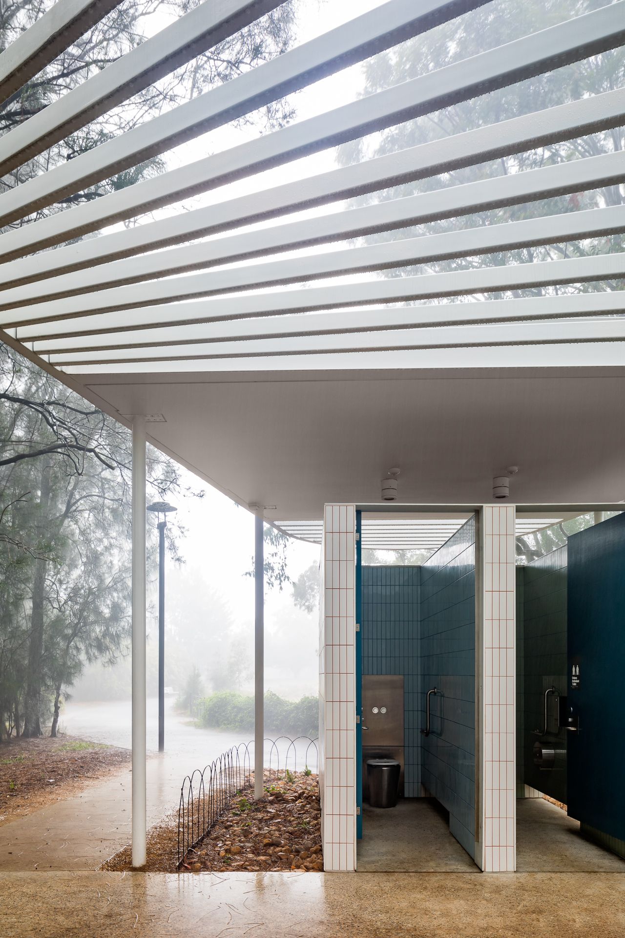 Modern park amenities block with white tiled walls, teal doors and a slatted pergola roof in misty bushland.