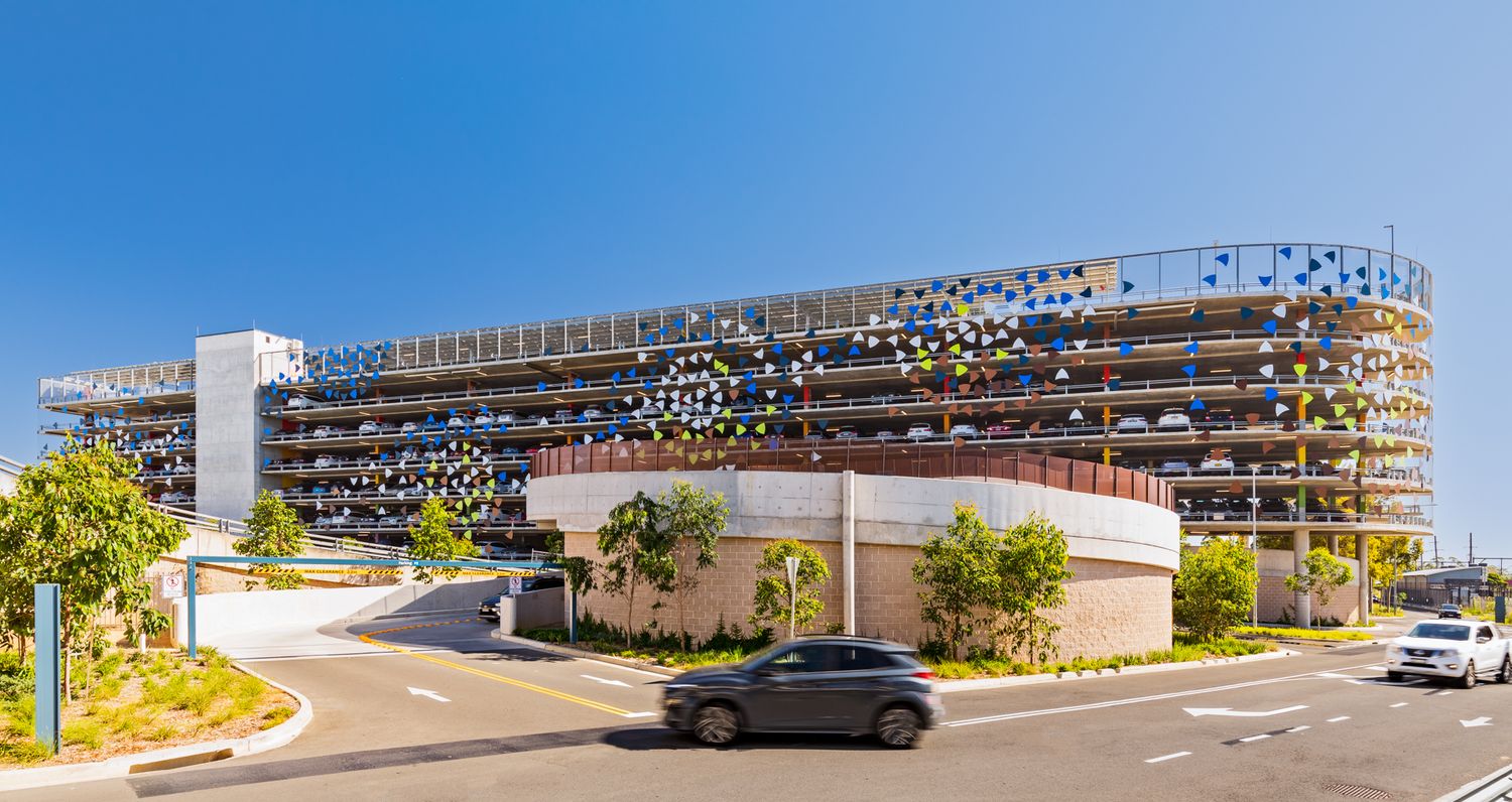 Modern multi-level car park with a colorful perforated facade, landscaped entrance and a car driving along the road.