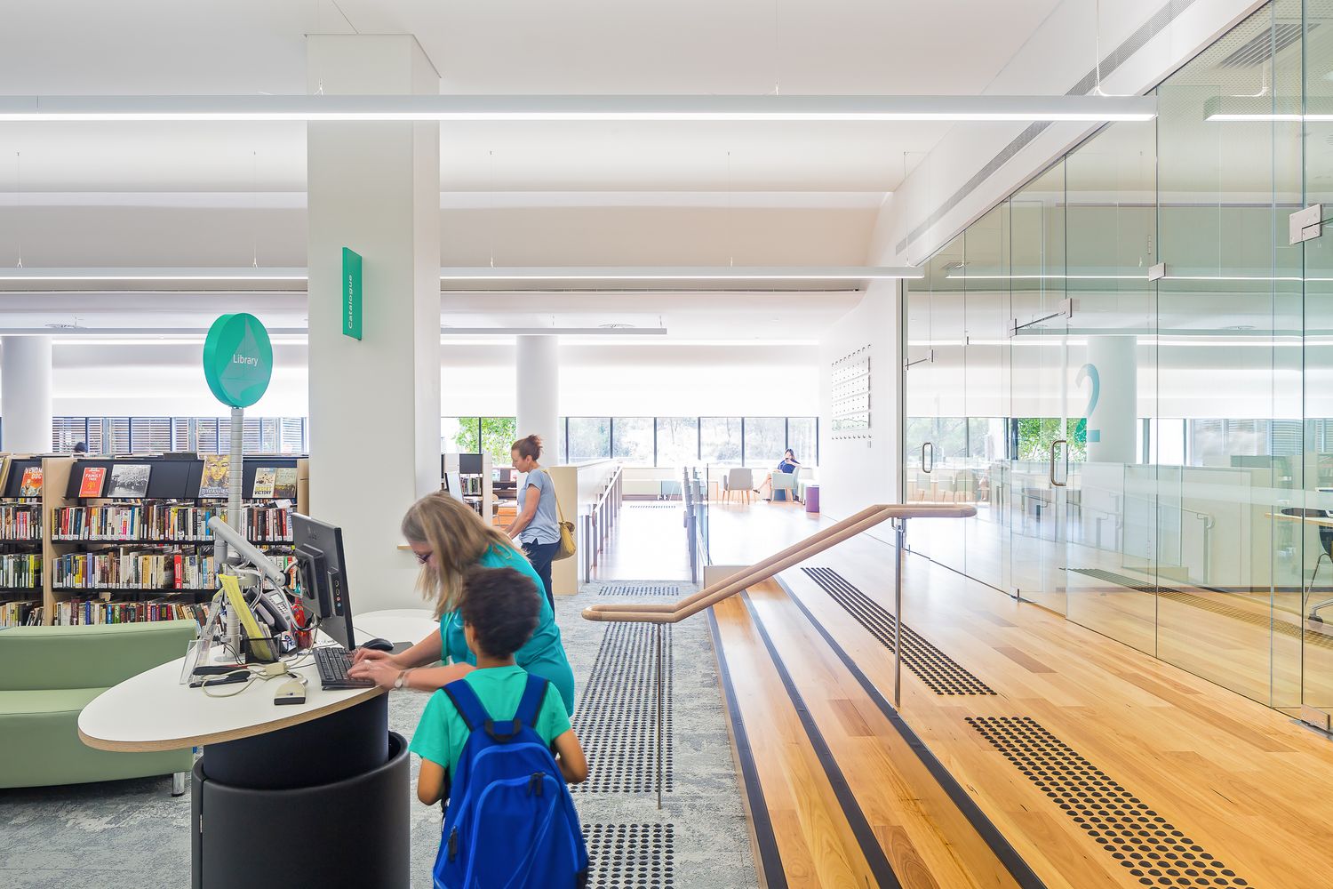 Bright modern library interior with timber stairs, bookshelves and a child with a backpack using a computer at a curved desk.