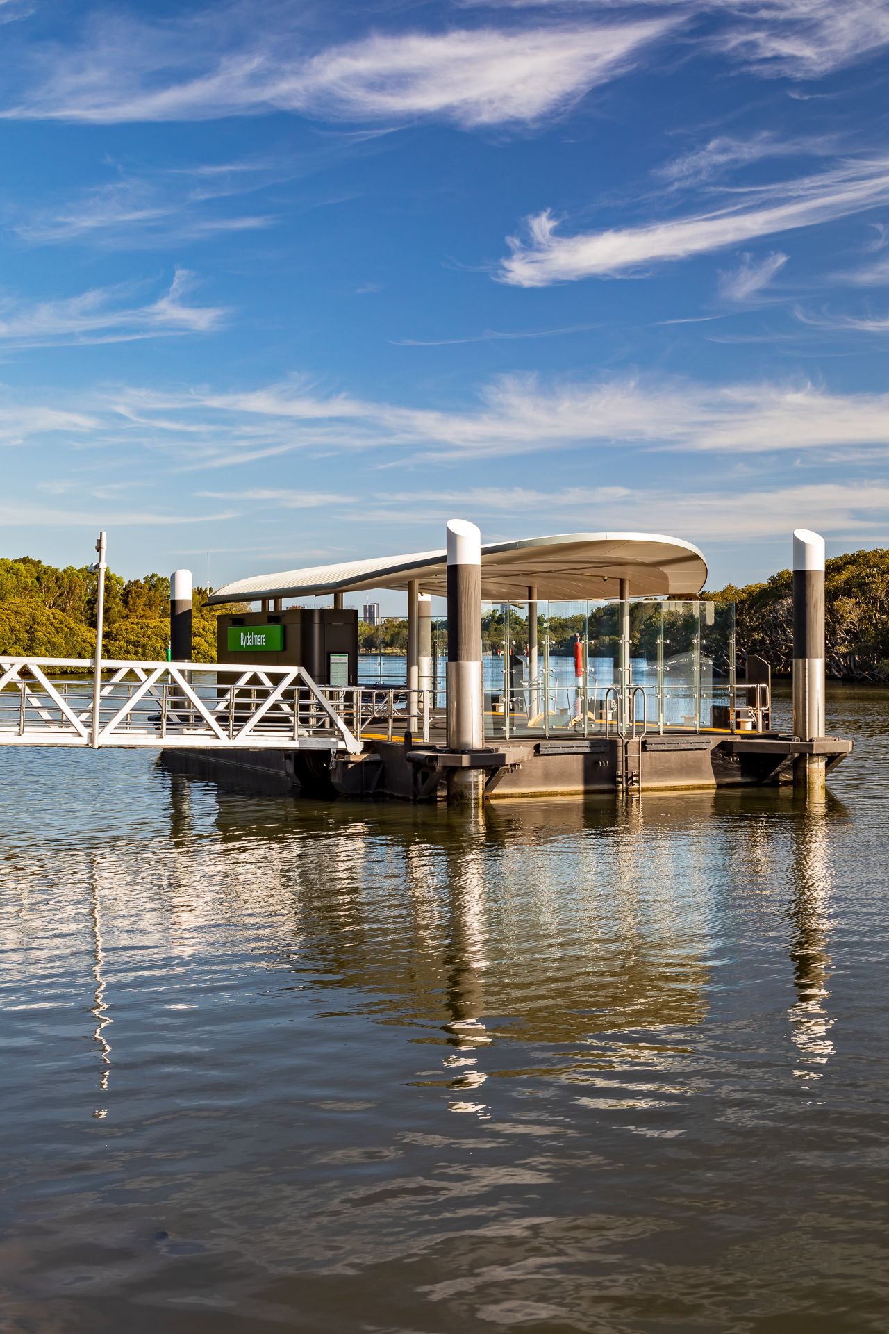 River ferry pontoon with a sleek canopy and white gangway reflected in calm water, with bushland and blue sky behind.
