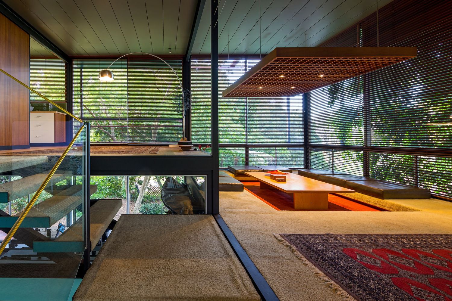 Modernist timber-lined living space with floor-to-ceiling glass walls, sunken seating area, red rug and views of surrounding bushland.