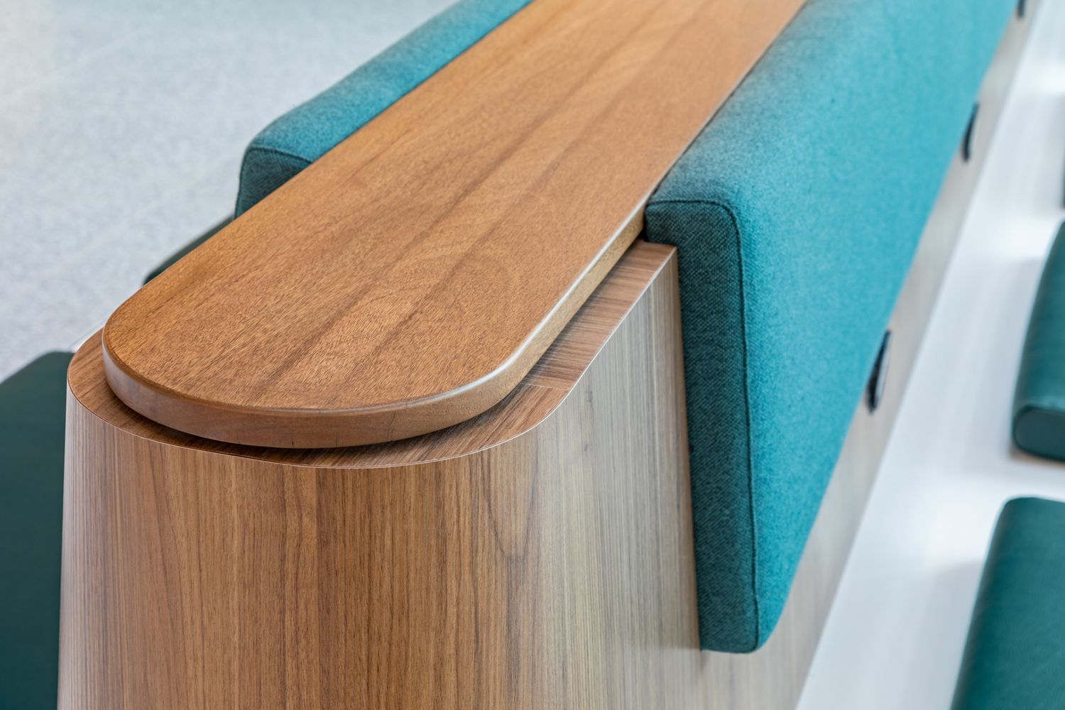 Close-up of a curved wooden armrest and teal upholstered seat backs in a modern lecture hall or seating area with warm wood paneling.