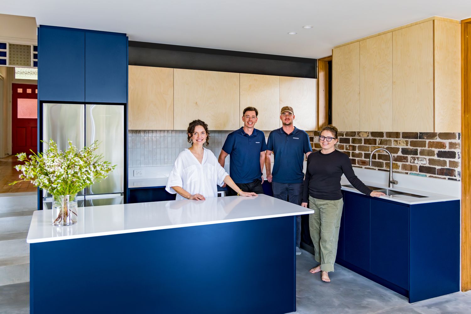 Four people smile in a newly renovated kitchen with navy blue cabinetry, white benchtops and a fresh flower arrangement.