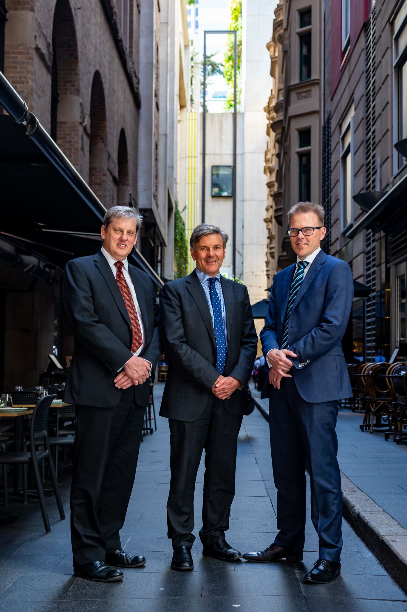 Three businessmen in suits and ties pose in a narrow city laneway lined with historic buildings and outdoor cafe tables.