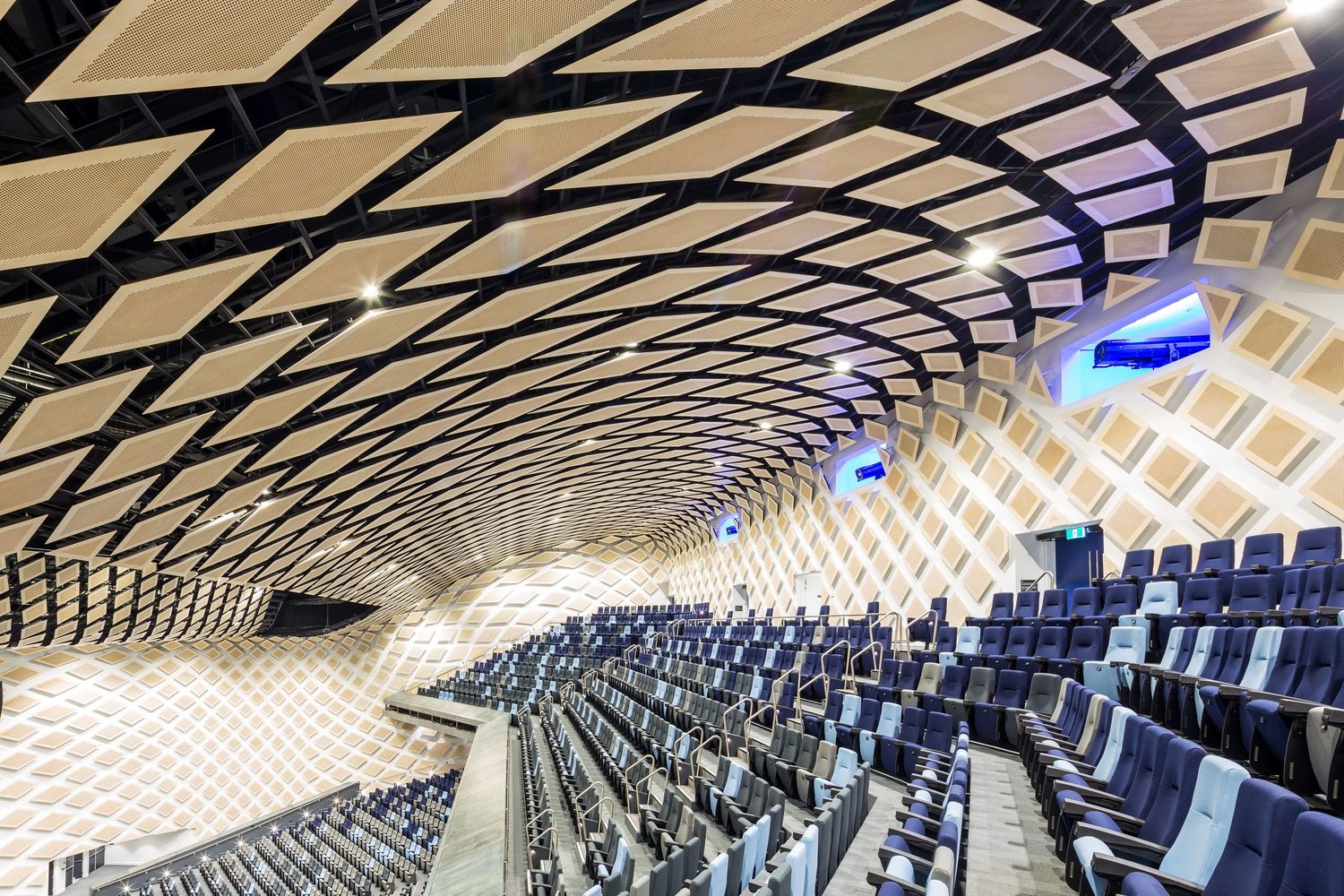 Curved lattice ceiling of a lecture theatre with warm timber panels, angled blue seating and illuminated screens.