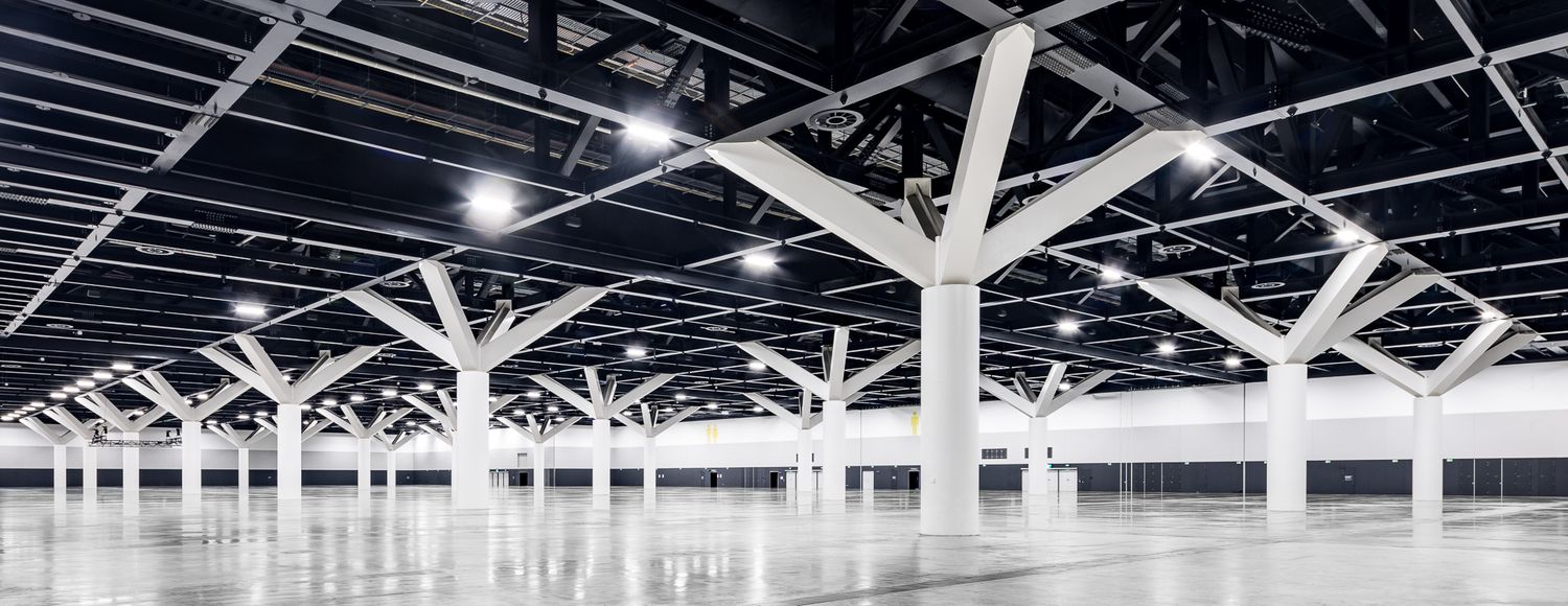 Expansive exhibition hall interior with polished concrete floors and rows of white branching steel columns under bright overhead lighting.