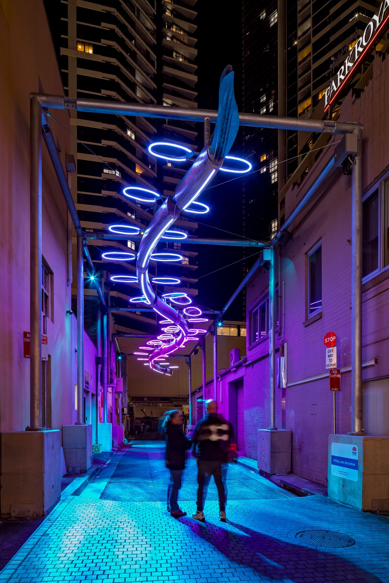 Two people pause in a blue-paved laneway under a looping purple and white neon light sculpture, with skyscrapers rising overhead at night.