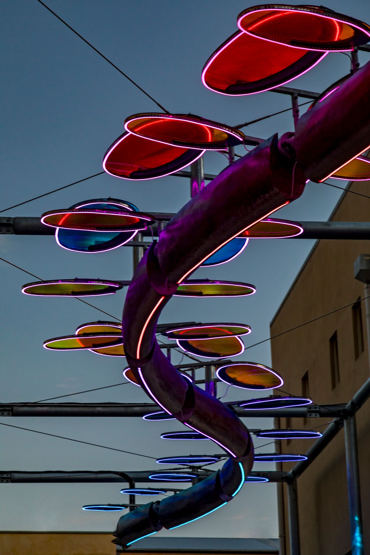Purple-lit serpent sculpture with pink-rimmed red and blue oval discs suspended on steel poles against a twilight sky.