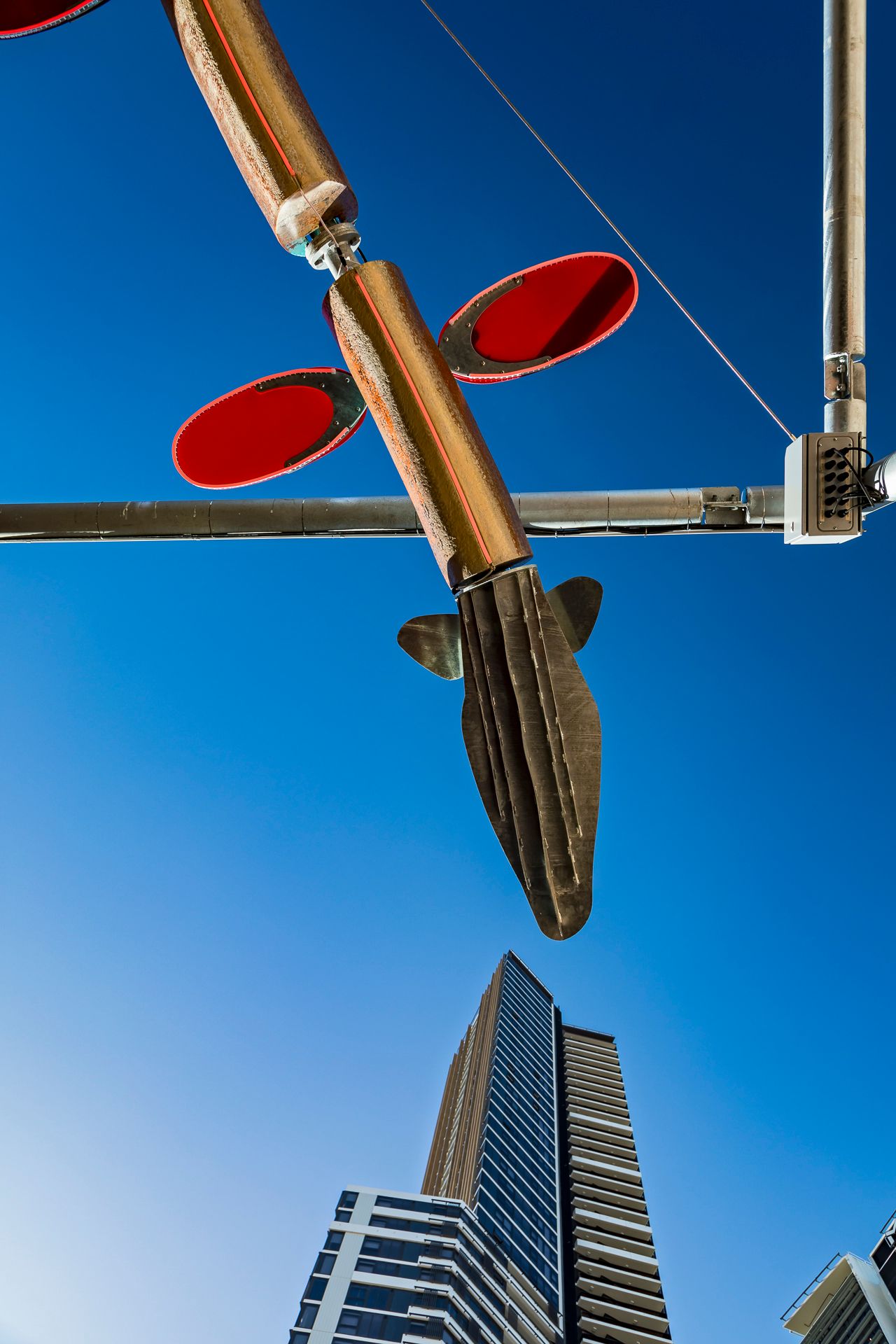 Fish-shaped metal tail and red oval discs of a suspended sculpture rise above a skyscraper in a clear blue sky.