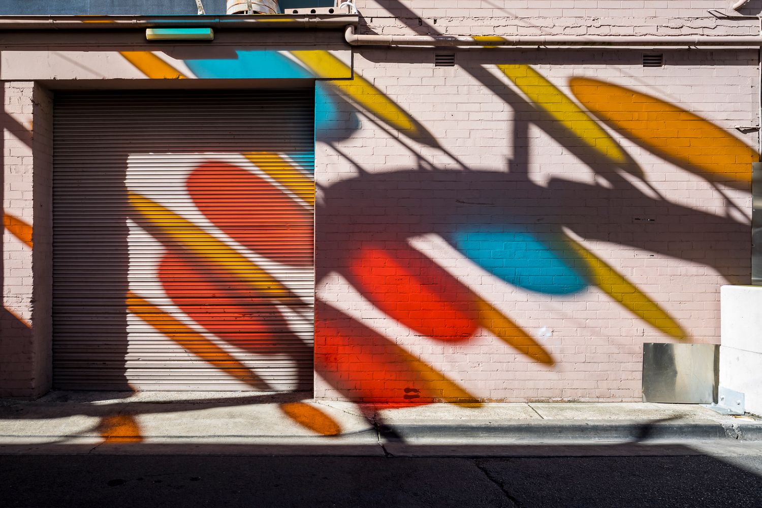 Vivid red, blue and yellow oval light patterns and shadows spread across a pink brick wall beside a closed roller shutter.