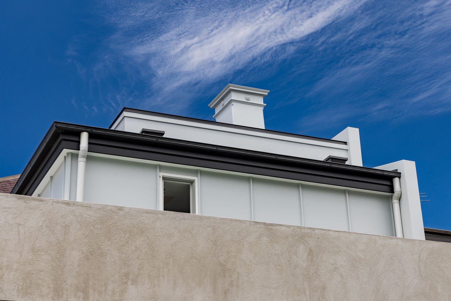 Upper exterior of a renovated period home with white walls, a dark roofline, a chimney and a concrete wall against a vivid blue sky.