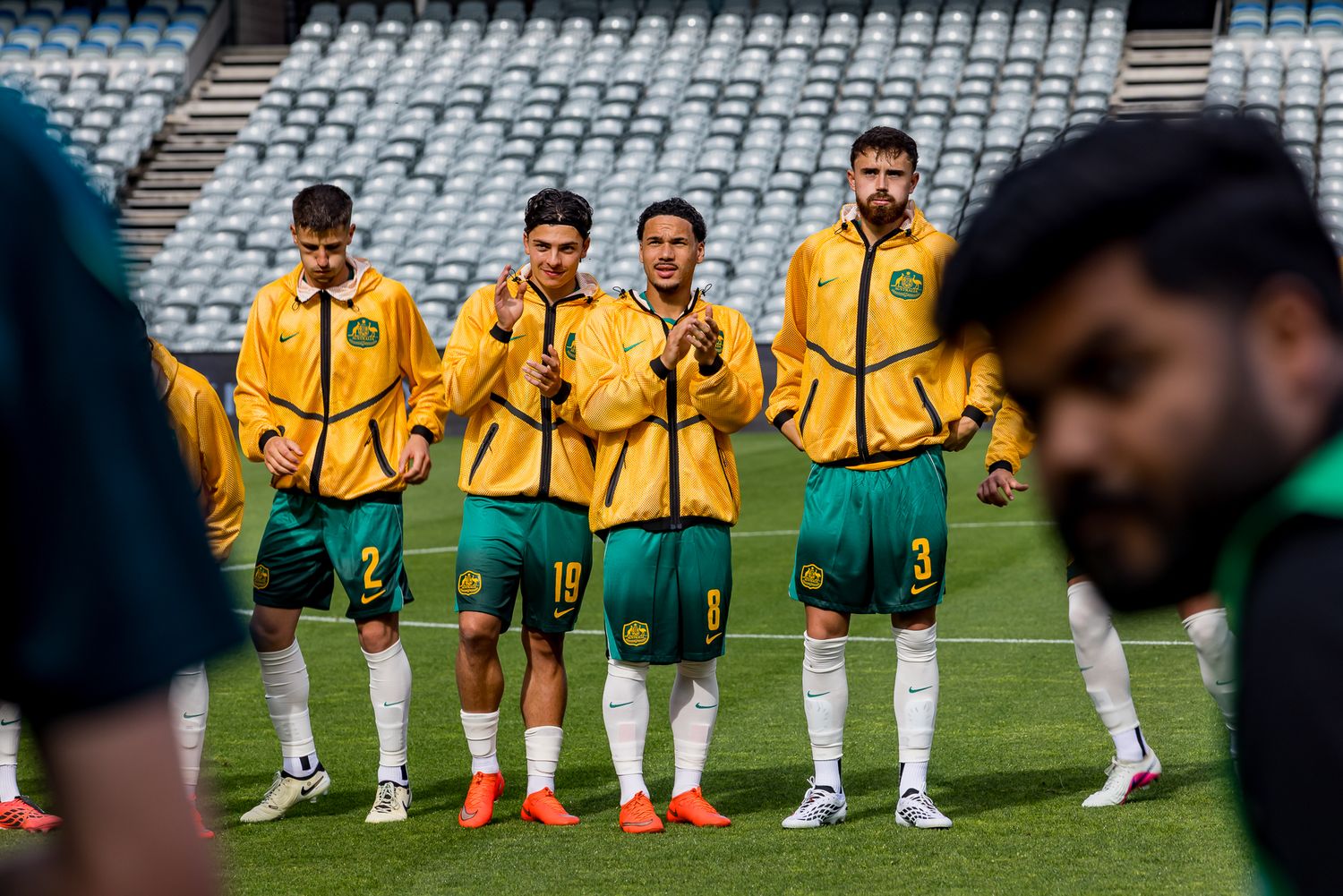 Australian soccer players in yellow training jackets and green shorts gather on the pitch ahead of a match.