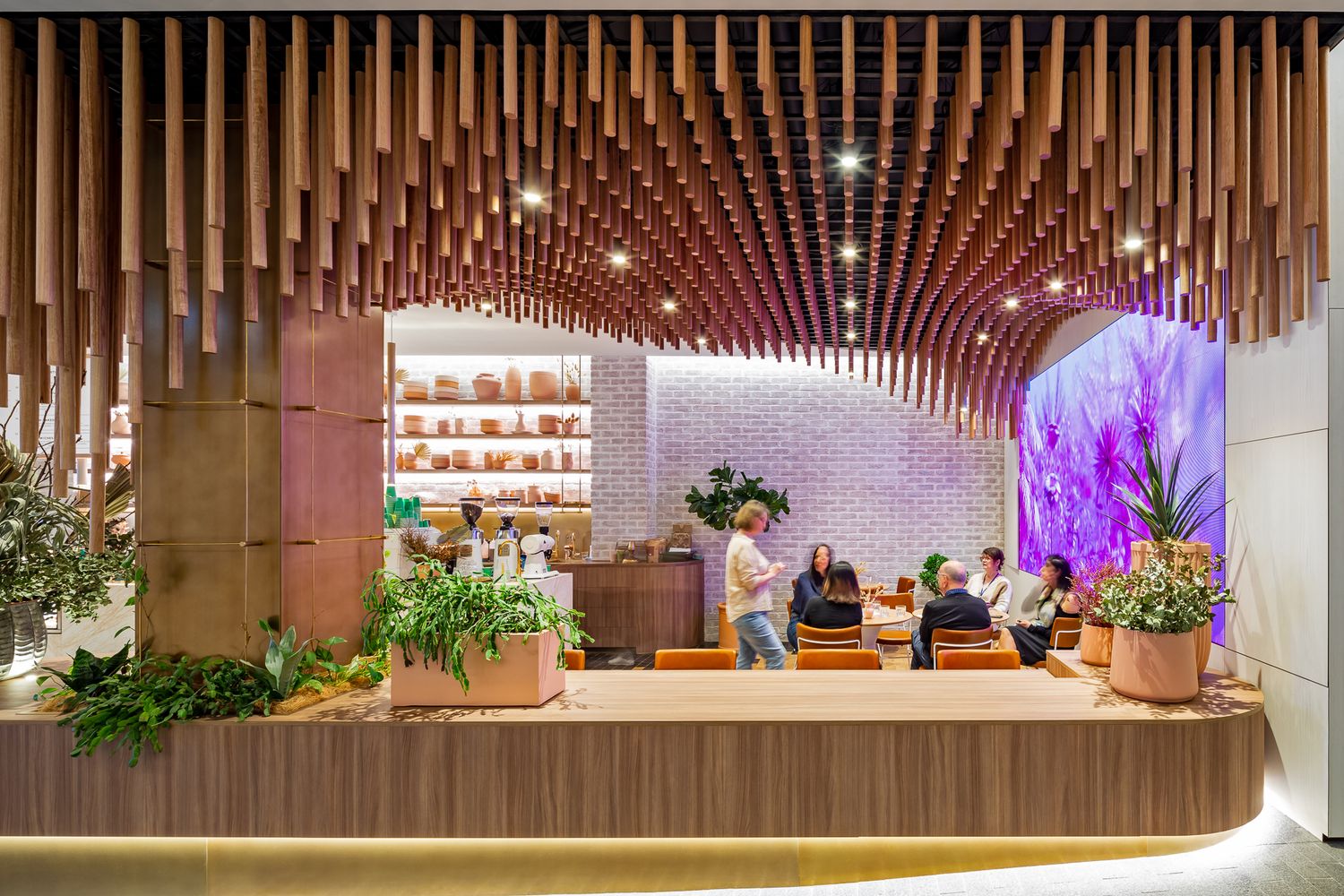 Cafe interior with customers seated at tables beneath a cascading timber tube ceiling, with a purple mural and potted plants.