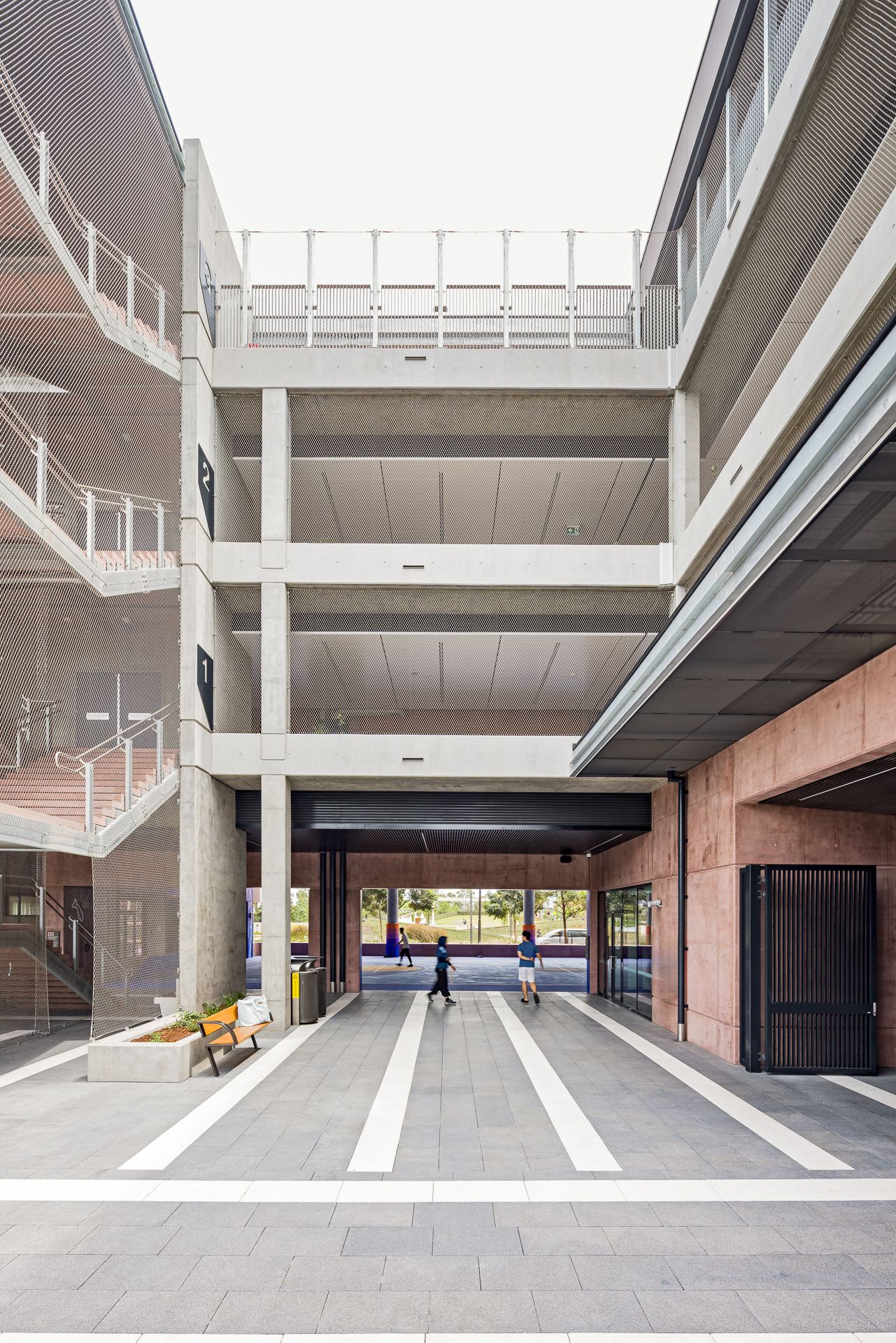 Open-air concrete courtyard with multi-level walkways, exposed columns and striped paving leading toward a landscaped street.