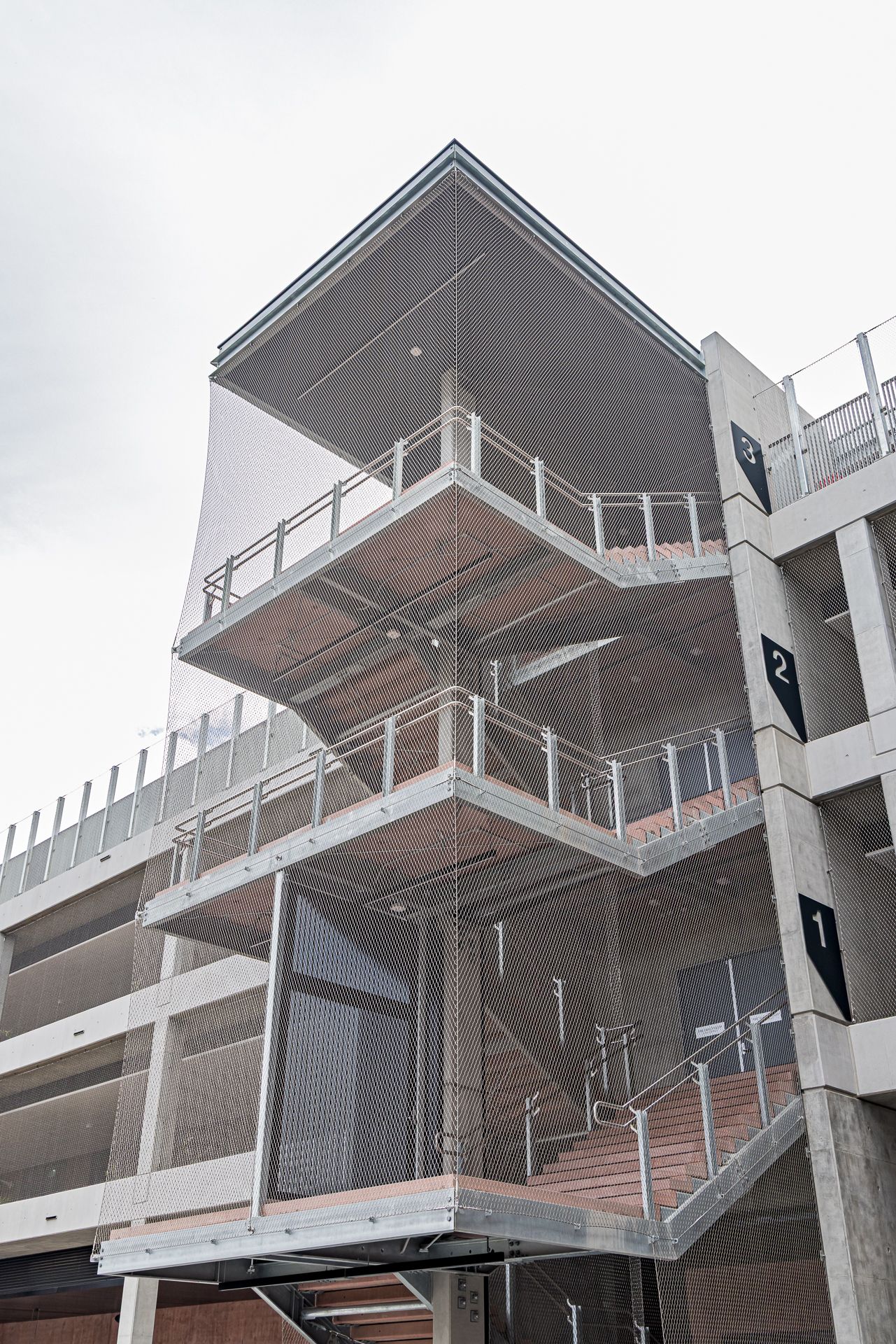 Open stair tower of a parking garage with numbered level signs, metal railings, mesh cladding, and concrete columns beneath a cloudy sky.
