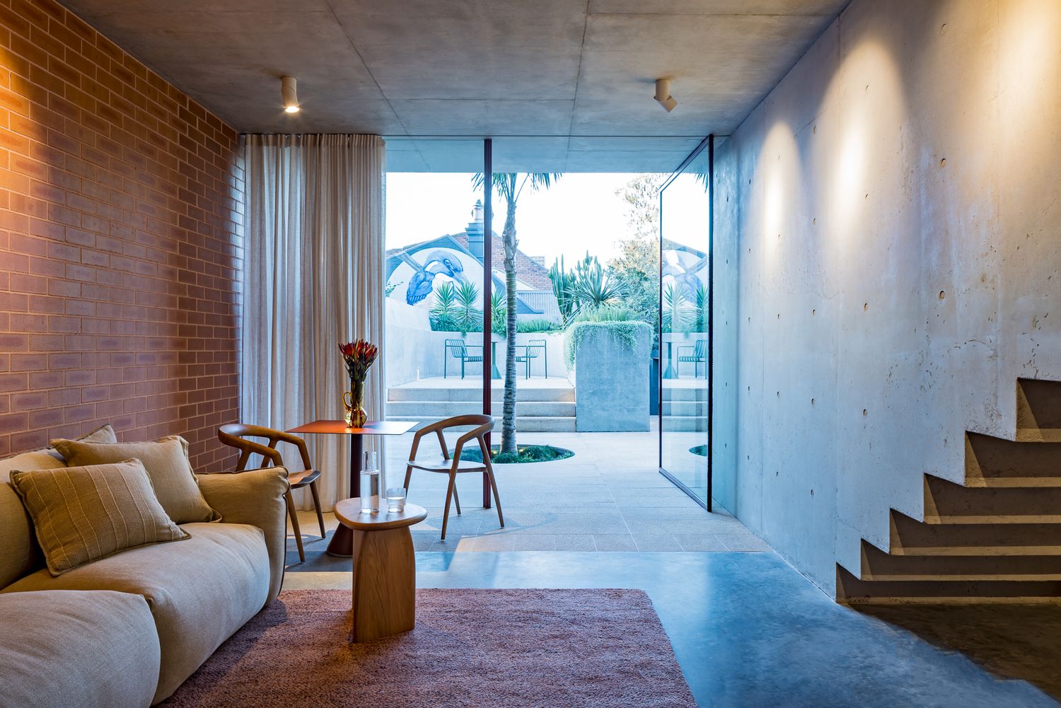 Contemporary lounge with brick wall, cream sofa, wooden chairs and concrete stairs overlooking a courtyard through glass walls.