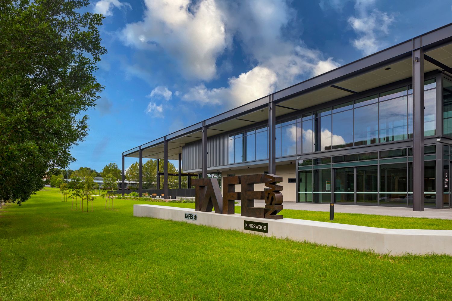 Modern TAFE NSW campus building with floor-to-ceiling glass and steel columns, large lettering sign, and green lawn under a blue sky.
