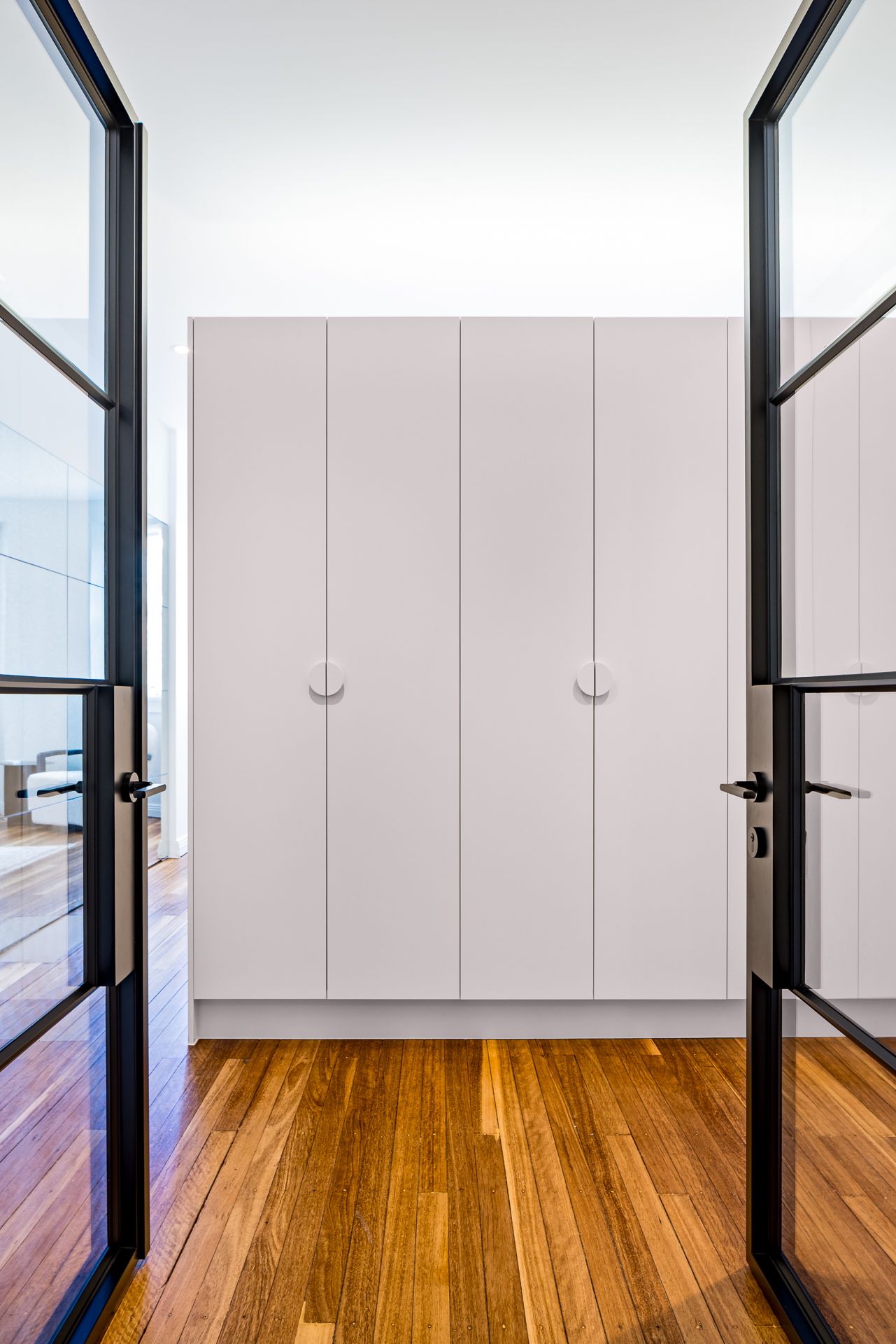 Black-framed glass doors open to a tall white built-in wardrobe with round handles and warm timber flooring.