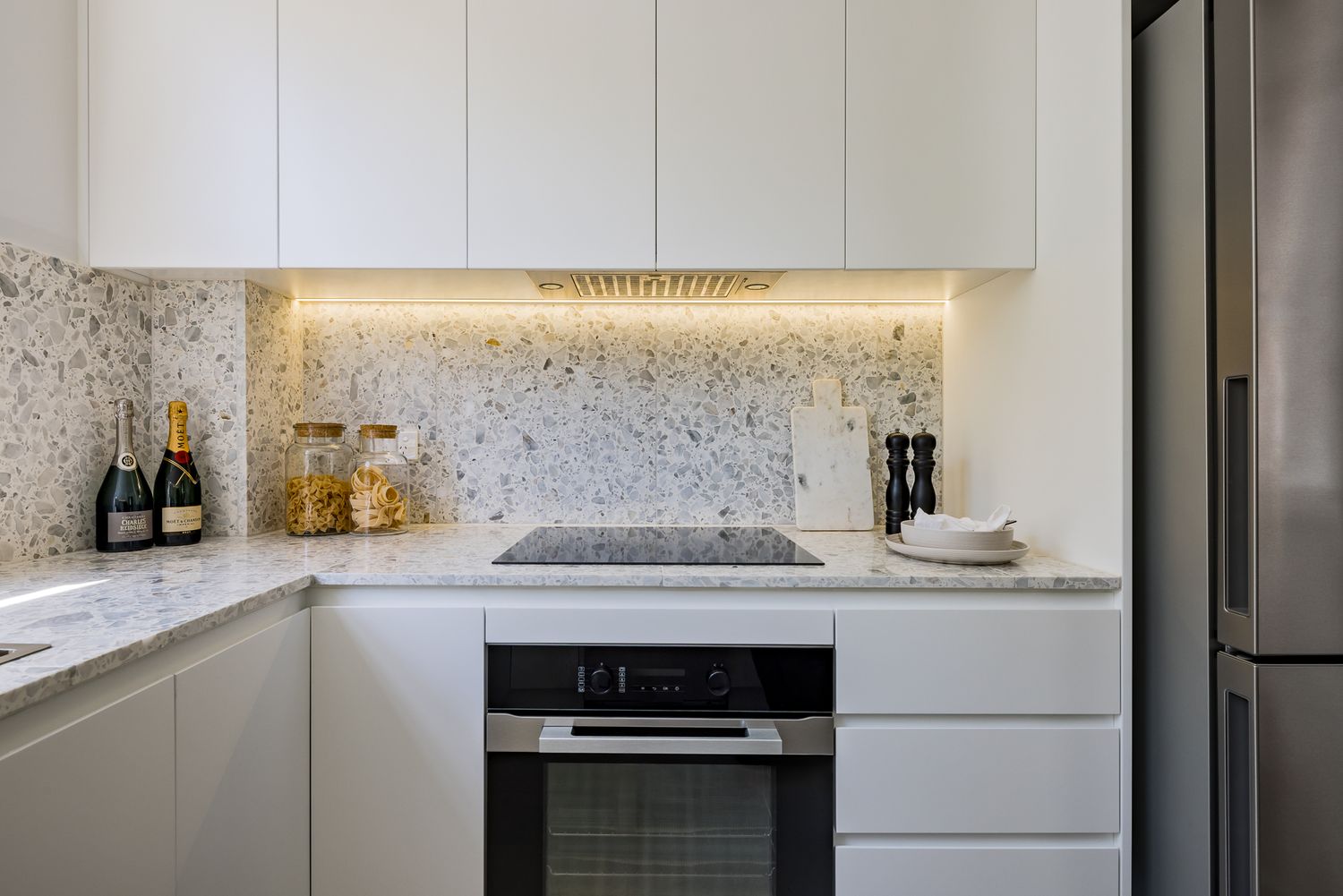 Contemporary kitchen with induction cooktop, built-in oven, white cabinetry and speckled stone splashback with under-cabinet lighting.