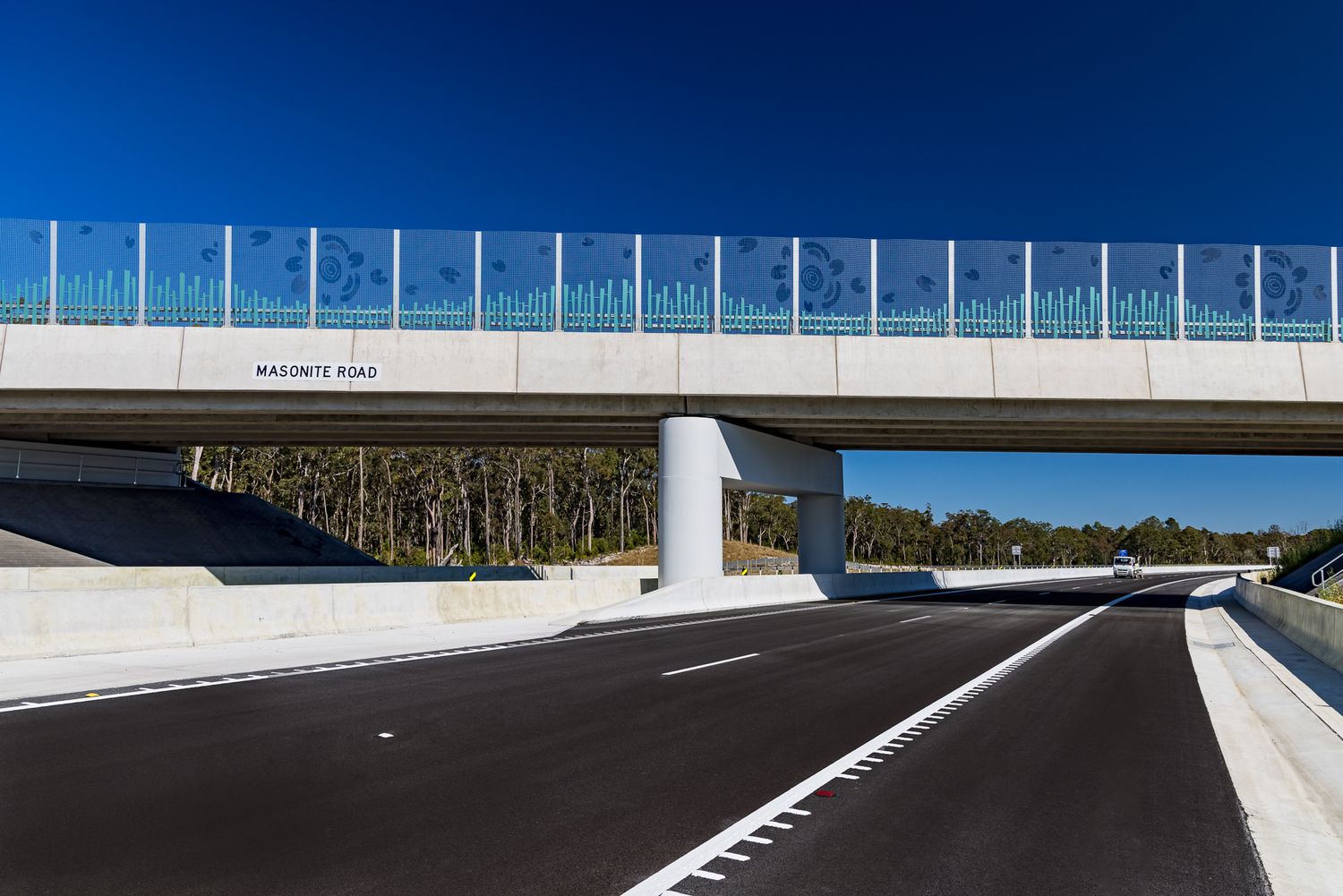 Modern concrete overpass with decorative blue glass panels spanning a freshly paved highway lined with trees under a deep blue sky.