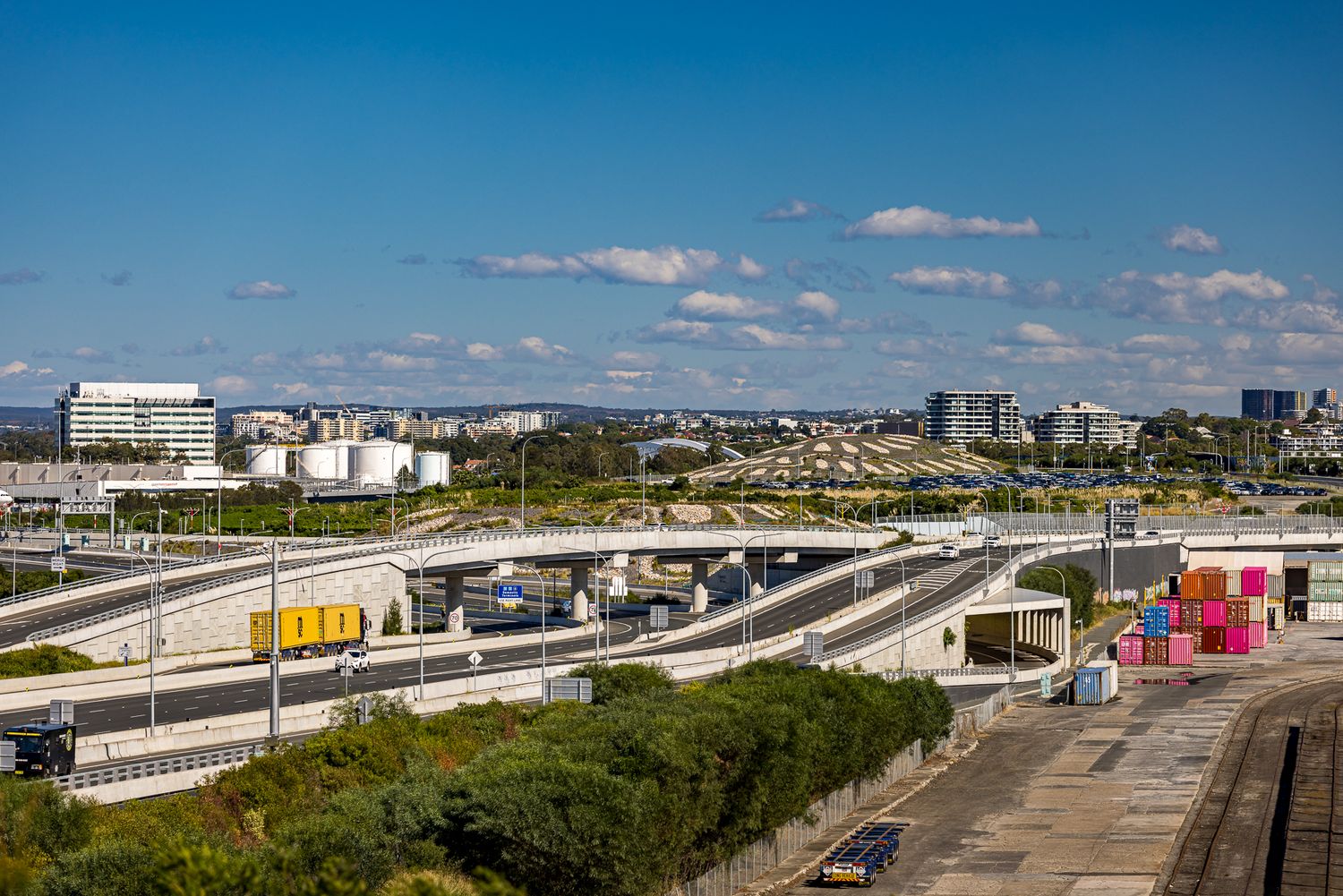 Elevated highway ramps and overpasses curve through an urban landscape with industrial buildings and a bright blue sky in the background.