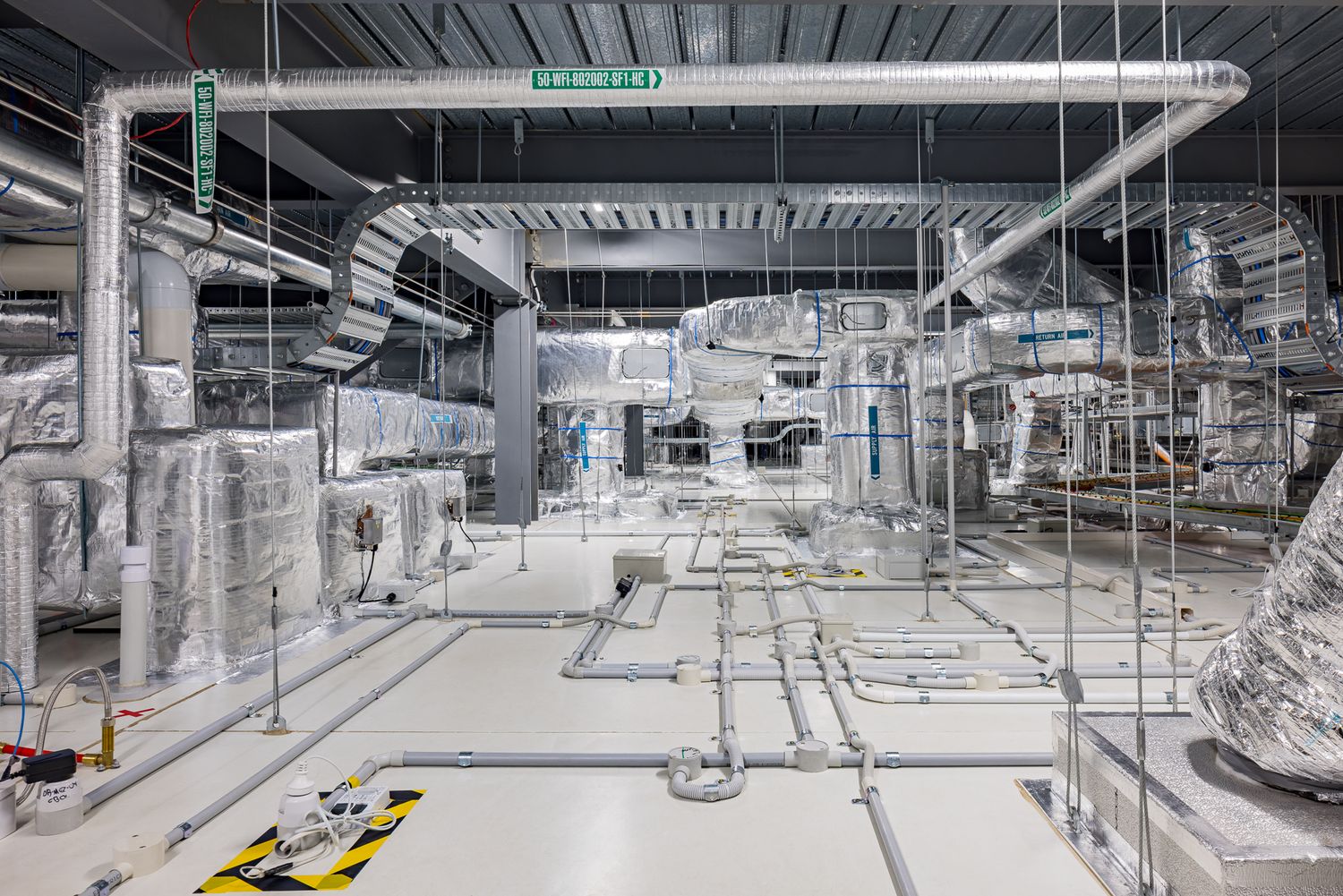 Large plant room with silver insulated ductwork, overhead pipes and a clean grey floor marked with yellow and black hazard tape.