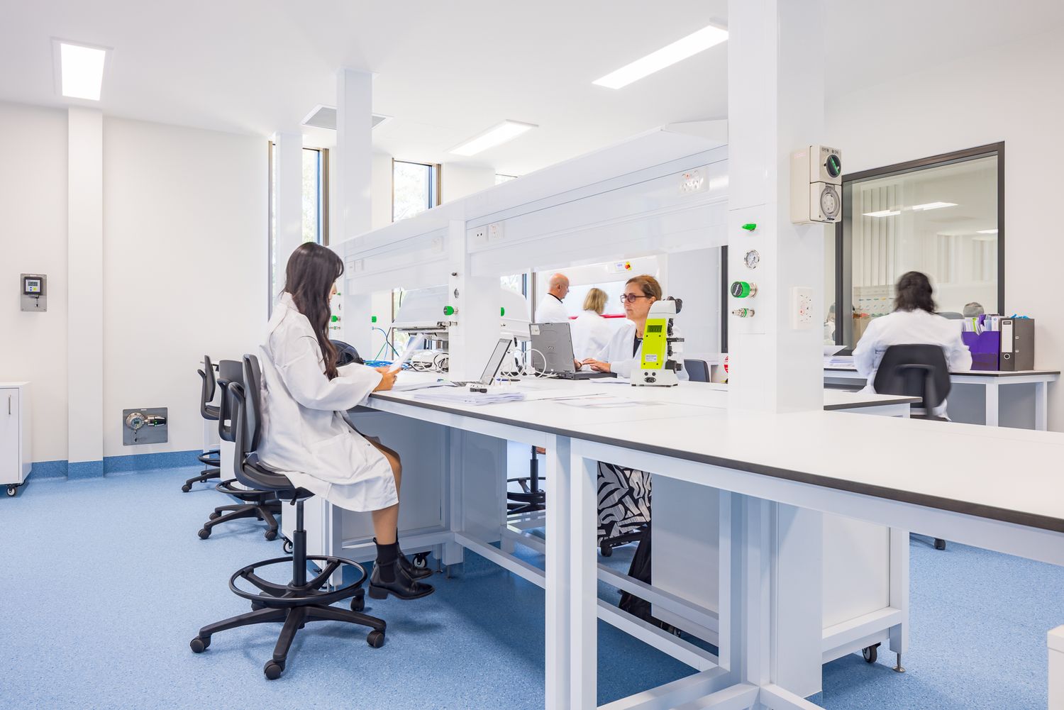 Researchers in white lab coats work at long white benches in a clean, bright laboratory with blue flooring and tall windows.