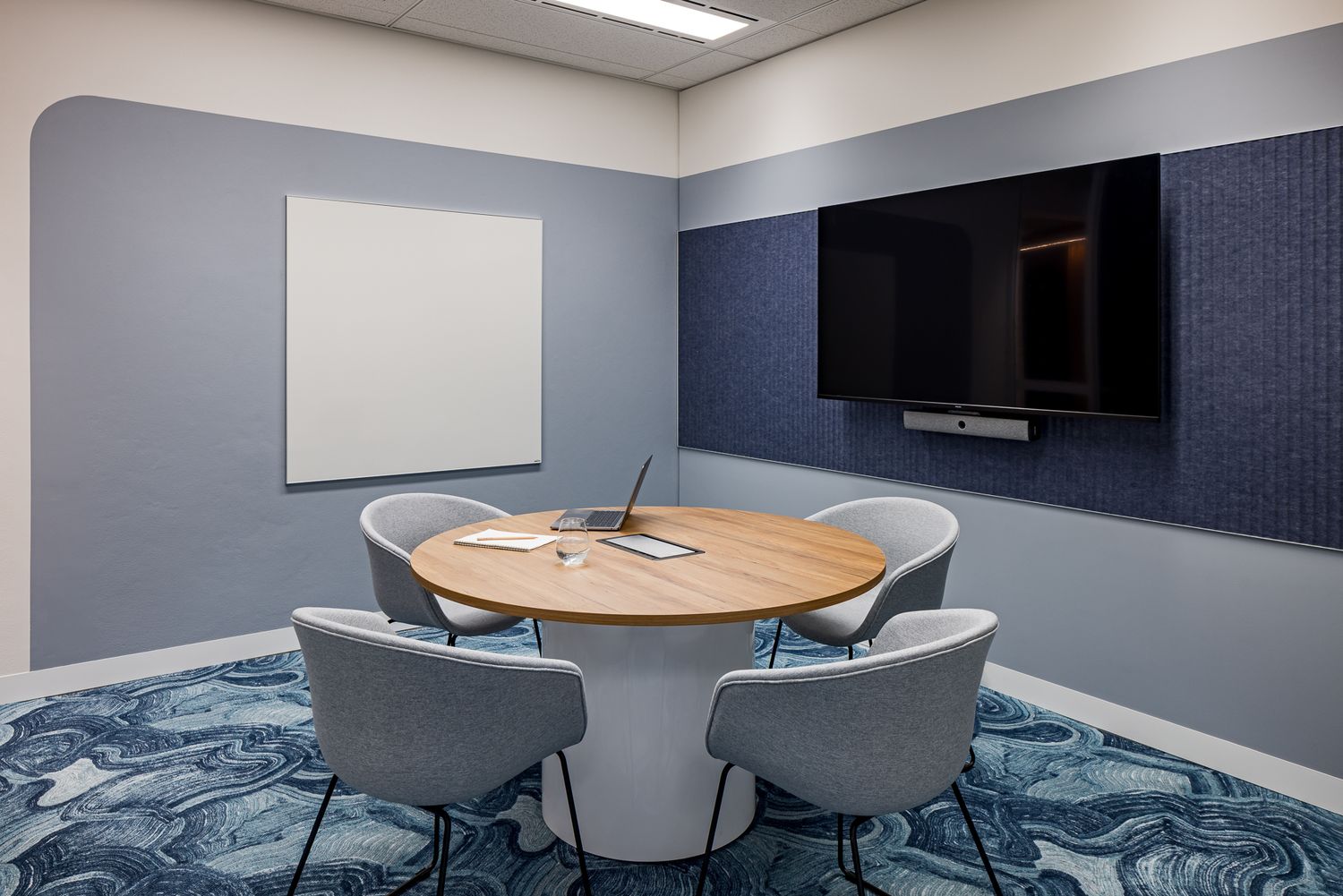 Compact meeting room with a round wooden table, gray upholstered chairs, wall-mounted TV, whiteboard and blue patterned carpet.