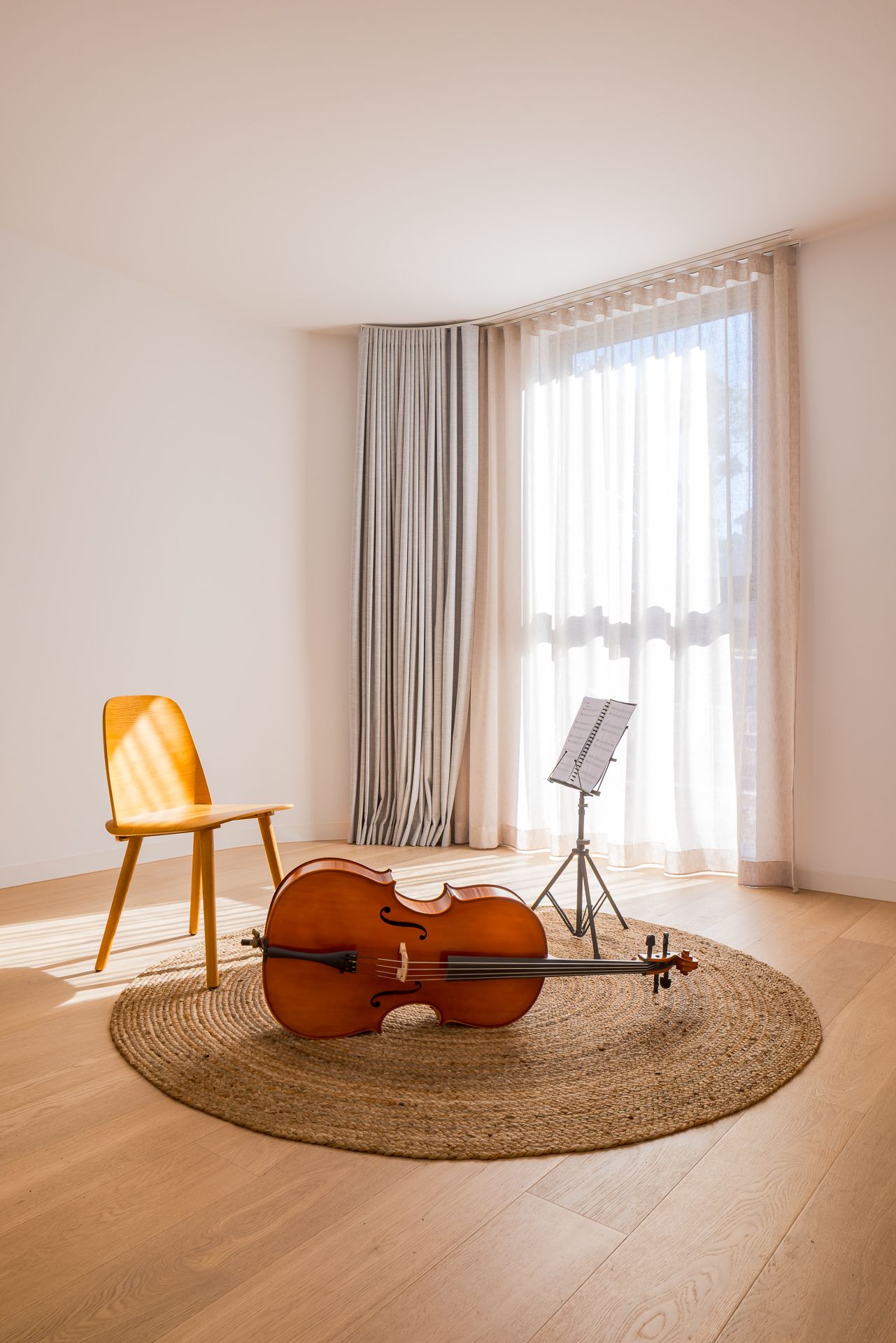 Bright minimalist room with a cello on a woven jute rug, a yellow wooden chair, a music stand and sheer curtains by a window.