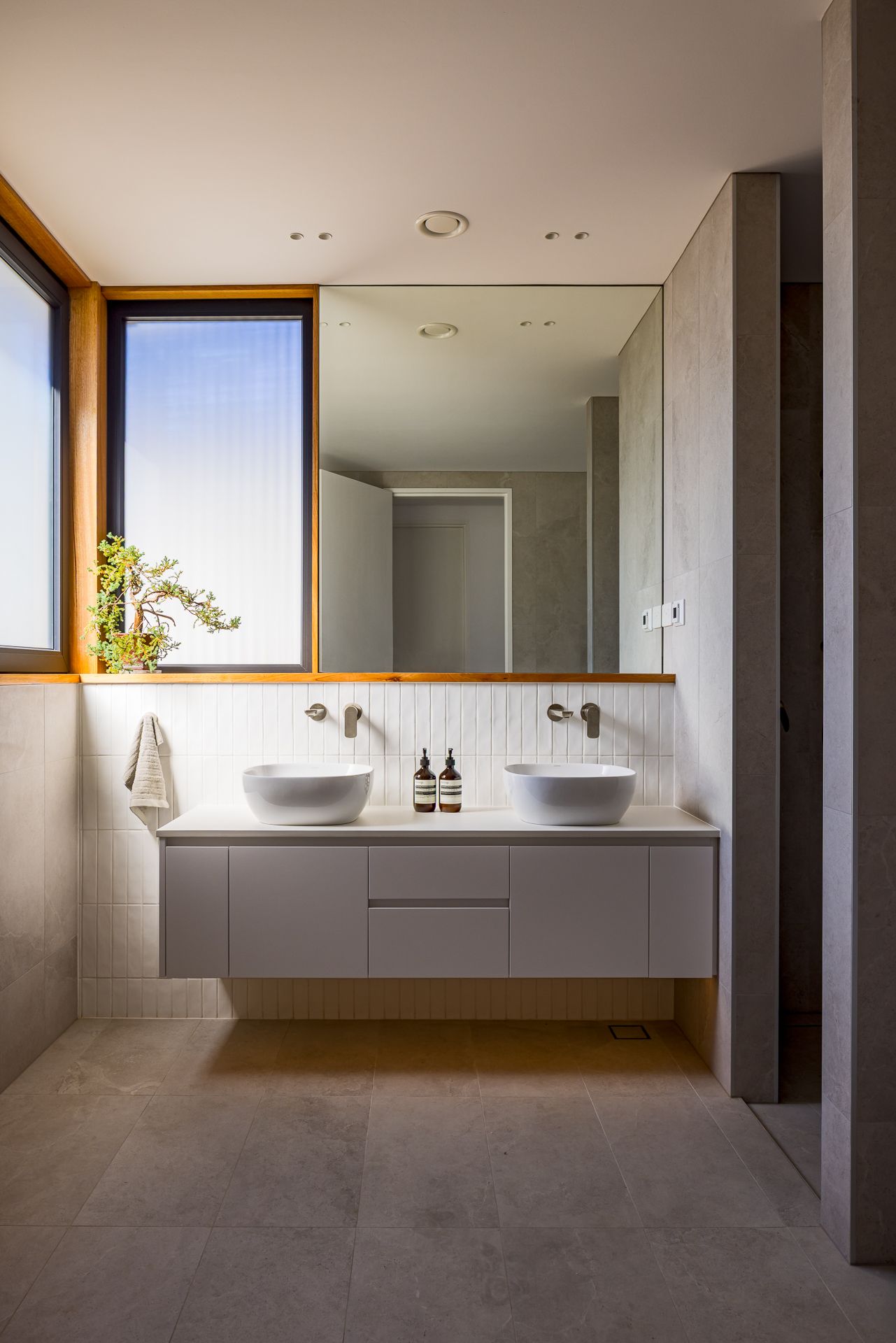 Serene bathroom featuring a wall-hung white vanity, two round basins, full-width mirror and warm timber window frame over stone tiles.