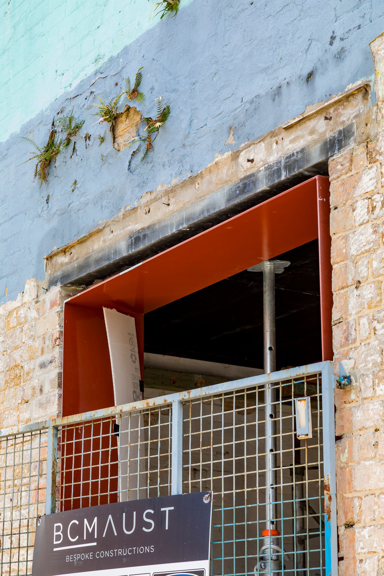 New rust-colored steel lintel framing a window opening in an old brick wall, with mesh security fencing and a BCMAUST sign.
