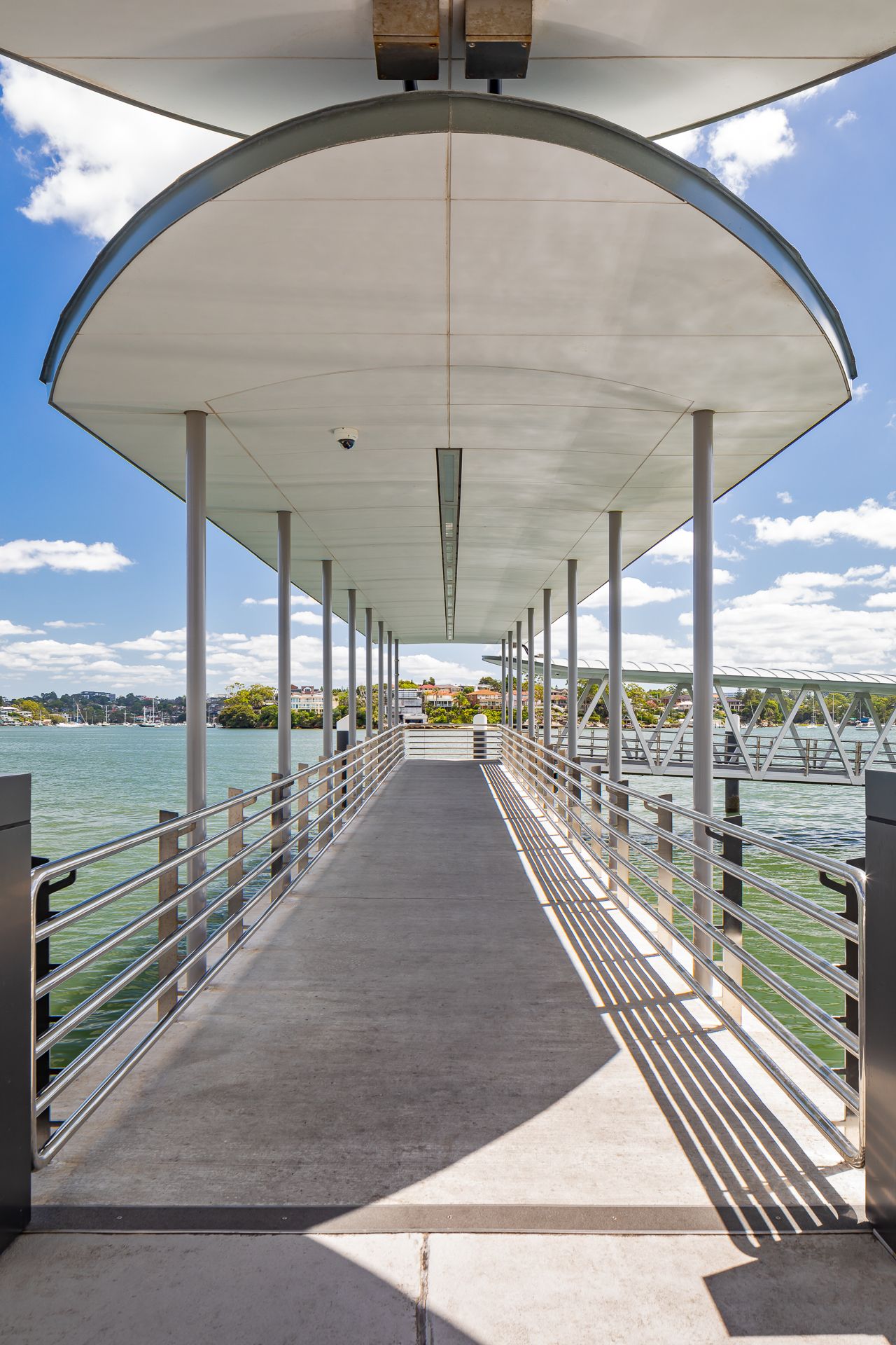 Covered ferry wharf walkway with metal railings and an arched roof leading over sparkling water under a blue sky.