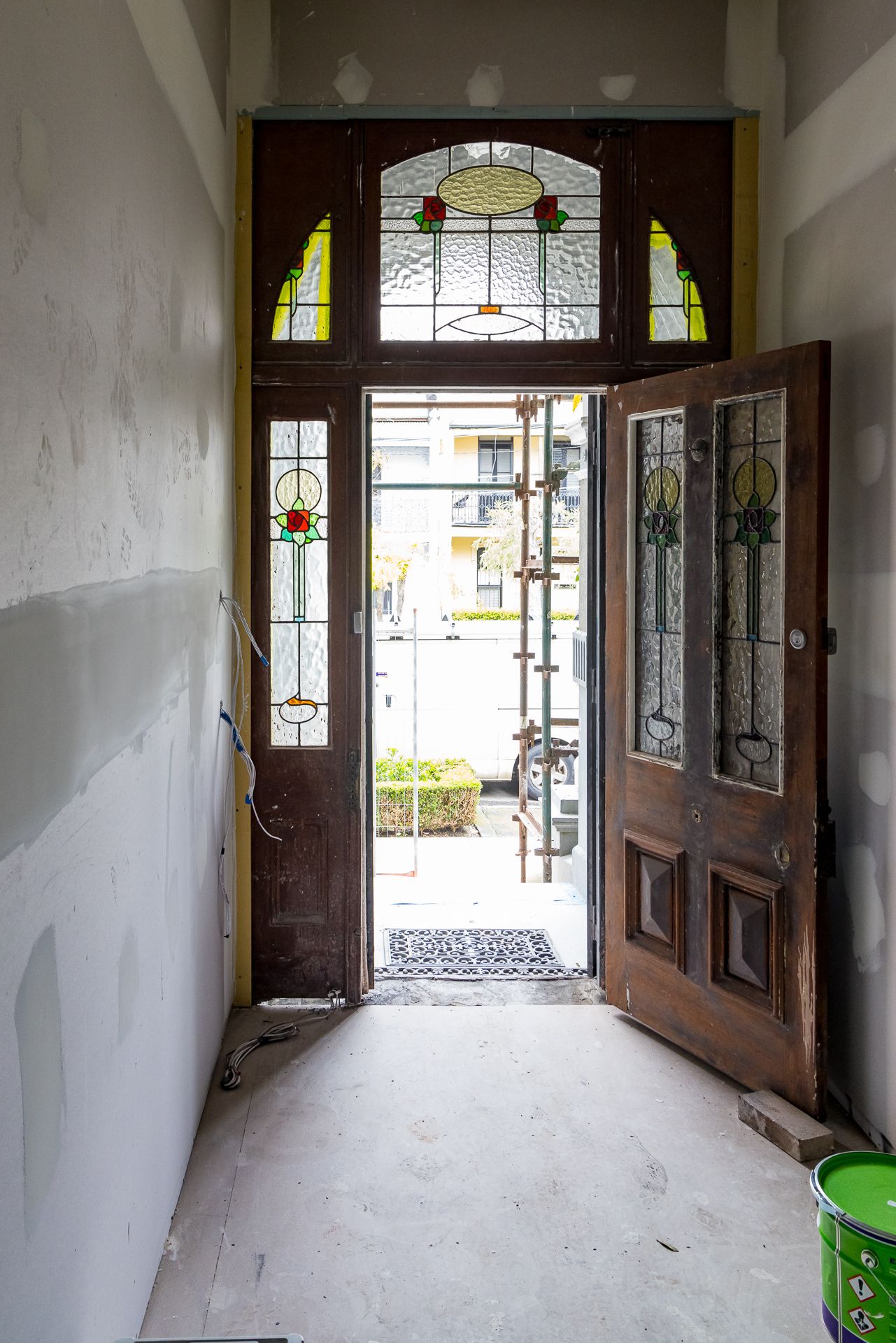 Open timber front door with stained glass panels and leadlight transom in a hallway with unfinished plastered walls.