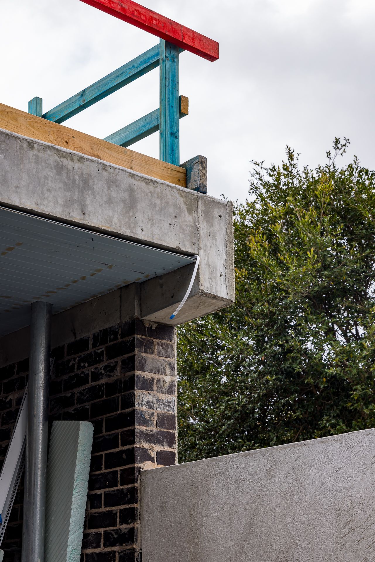 Concrete balcony edge with temporary blue and red safety railing above a dark brick wall and a leafy green tree at a construction site.