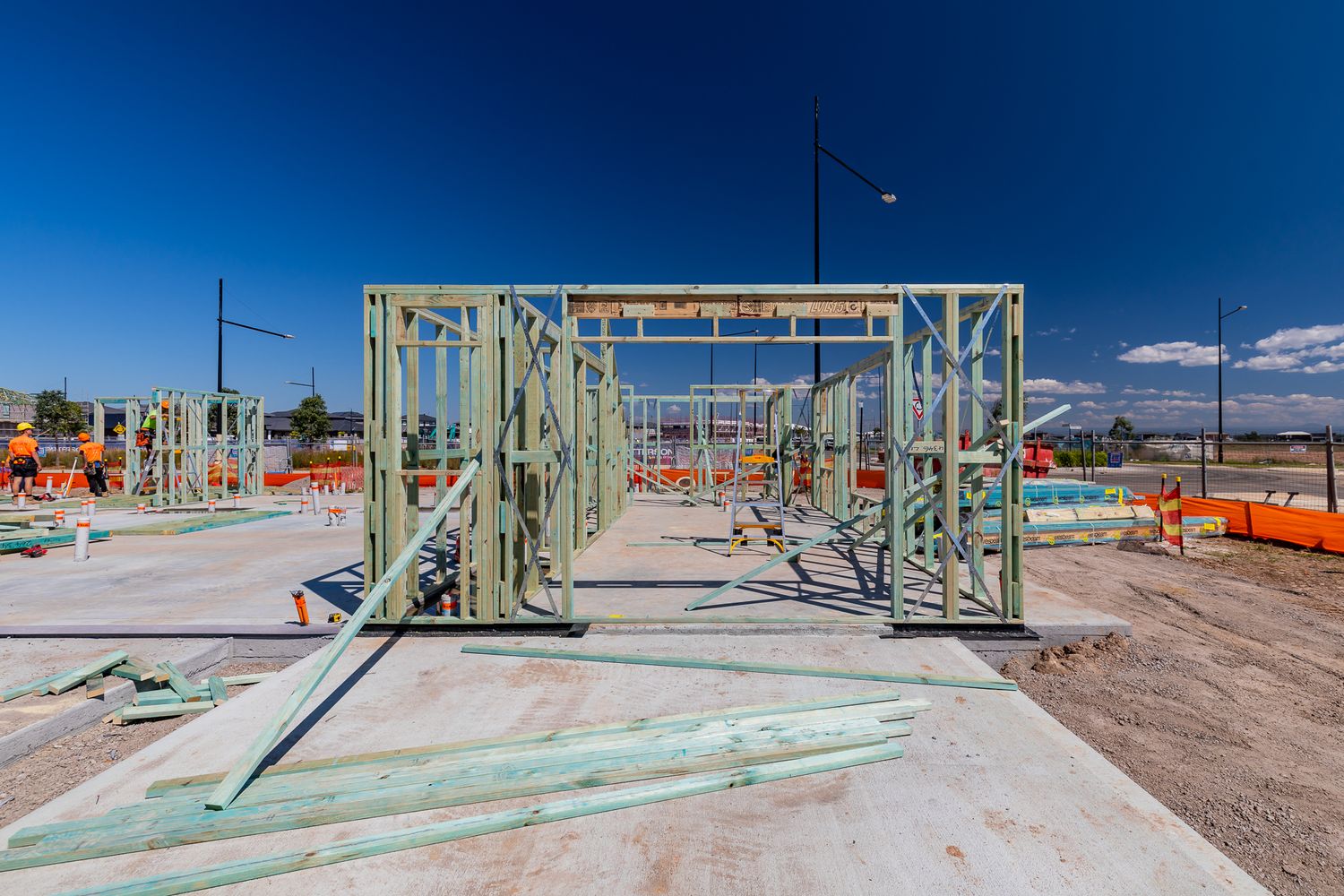 Timber and steel wall framing stands on a concrete slab at a residential building site under a clear blue sky.