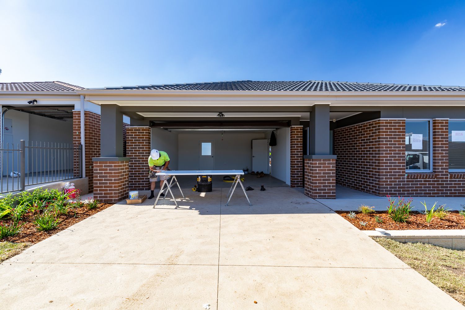 Newly built brick home with a covered porch, concrete driveway and a trestle table, with fresh landscaping beds along the front.