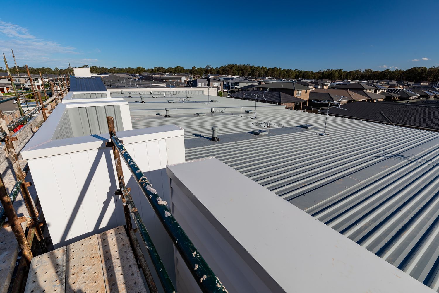 Rooftop view of a new townhouse development showing corrugated metal roofing, parapet walls and surrounding suburban homes.