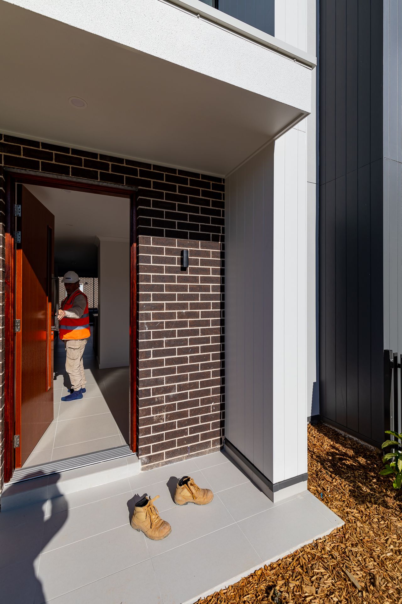 Builder in a hi-vis vest stands inside the open front door of a new brick townhouse, with work boots left on the entry porch.