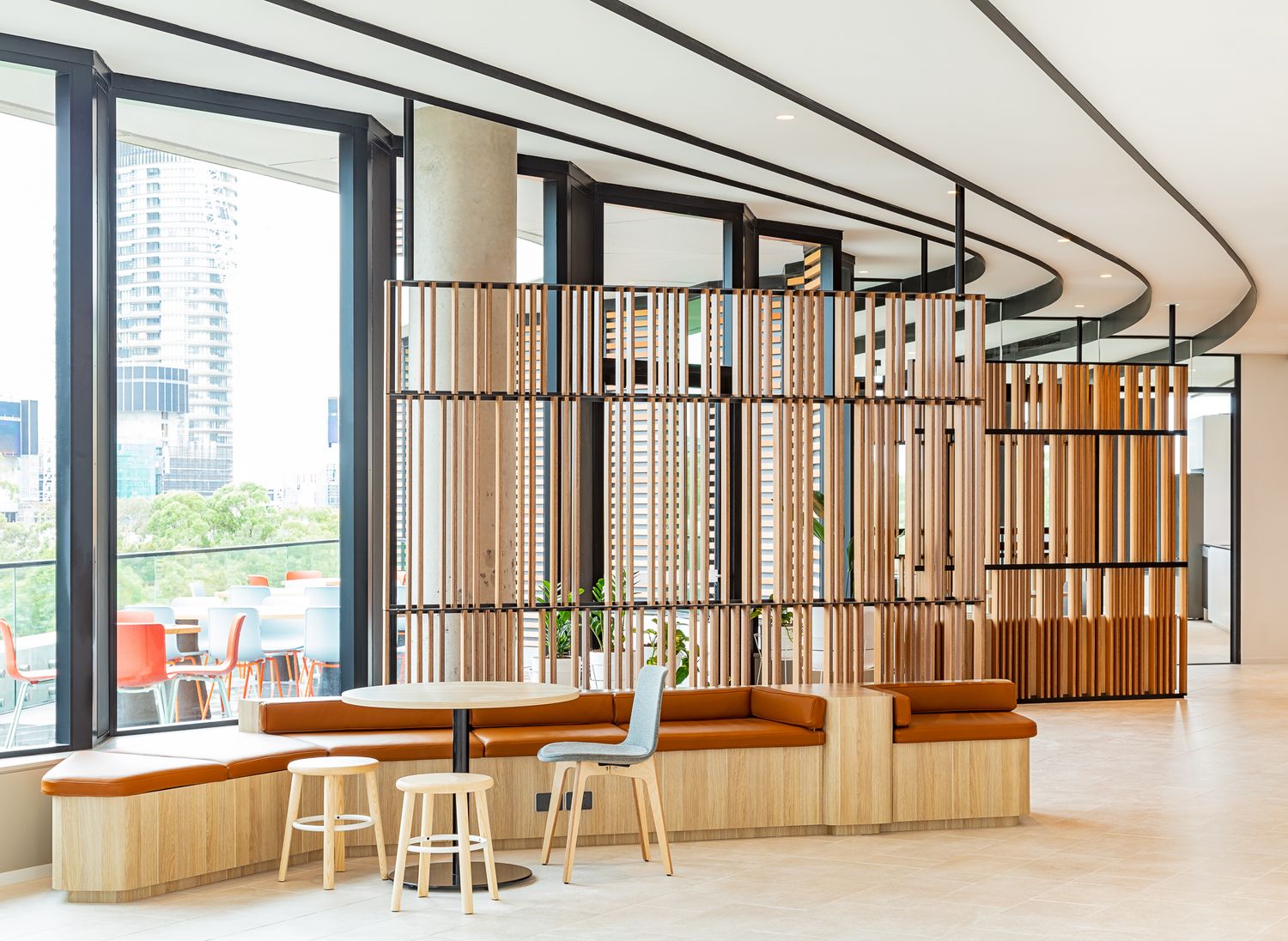 Modern office lounge with wooden slat dividers, curved banquette seating, bar stools and floor-to-ceiling windows overlooking the city.