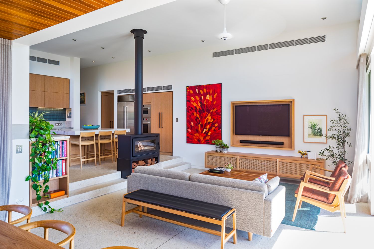 Bright open-plan living room with a freestanding black fireplace, grey sofa, timber cabinetry and a vivid red artwork.