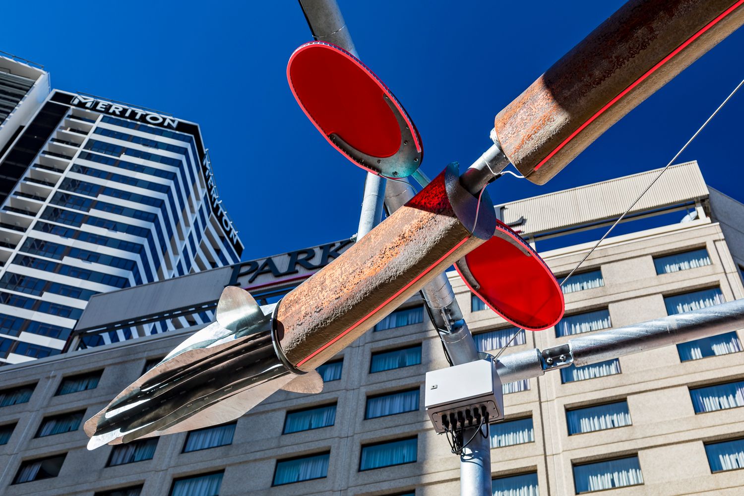 Bronze fish sculpture with red disc lights suspended over a city street, with hotel and apartment buildings in the background.