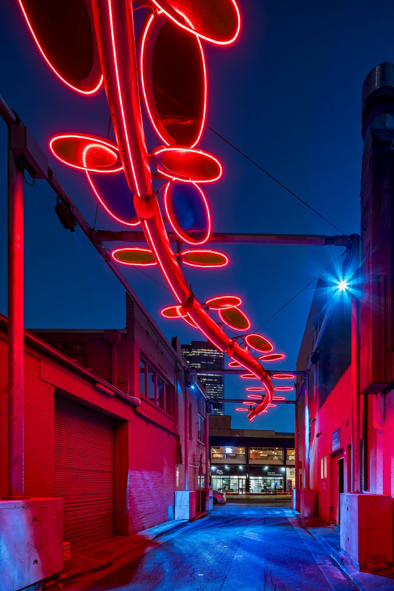 Vivid red neon oval ring sculpture suspended above a narrow city alley at night, bathing the brick walls in red light.