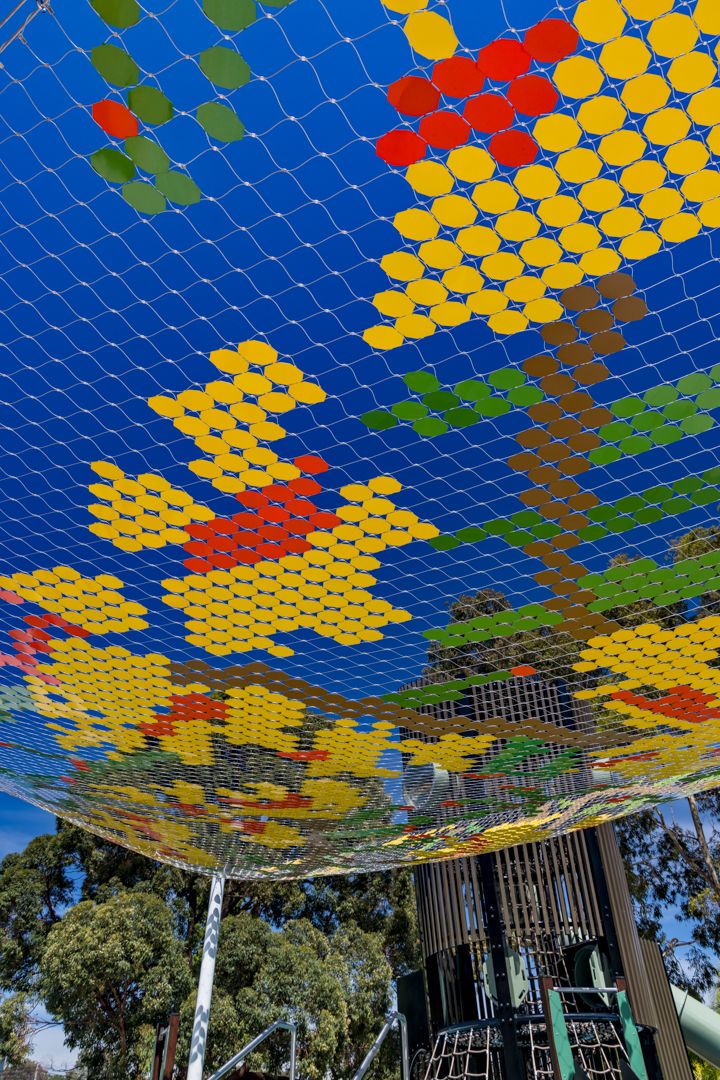 A colorful netted canopy with a pixelated flower and tree design stretches above a playground against a vivid blue sky.