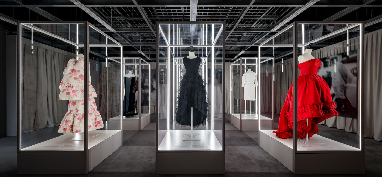 Fashion exhibition gallery with illuminated glass display cases showing pink, black and red couture gowns on mannequins.
