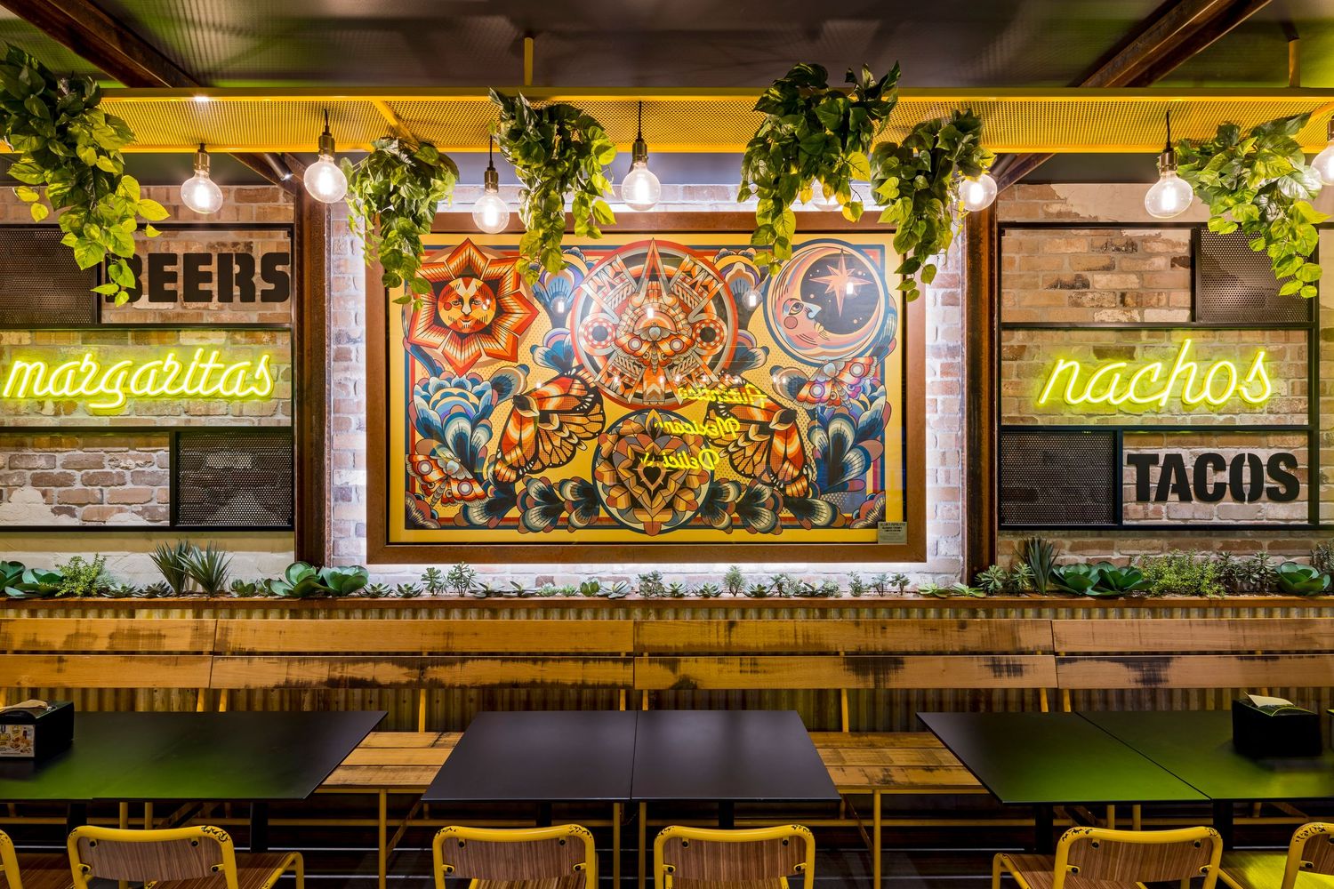 Restaurant interior with neon margaritas, nachos and tacos signs, hanging plants, wooden tables, and a colorful ornate framed mural.