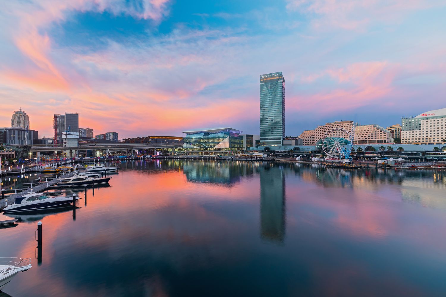 Sydney harbour at sunset with pink and orange clouds reflecting on still water beside waterfront buildings.