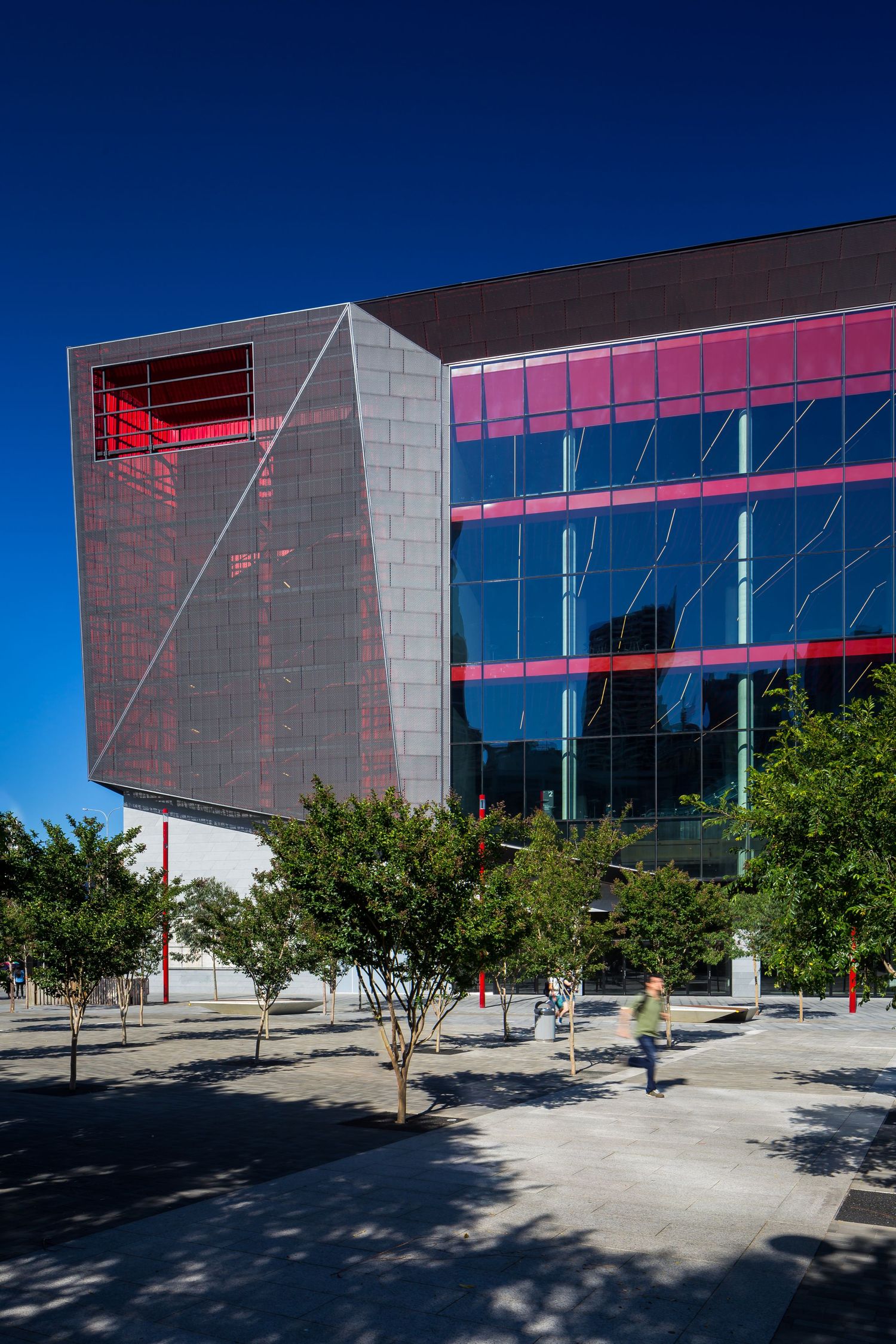 Striking convention center exterior with metallic angular panels, pink-tinted glass and a pedestrian walking beneath trees on a sunny plaza.